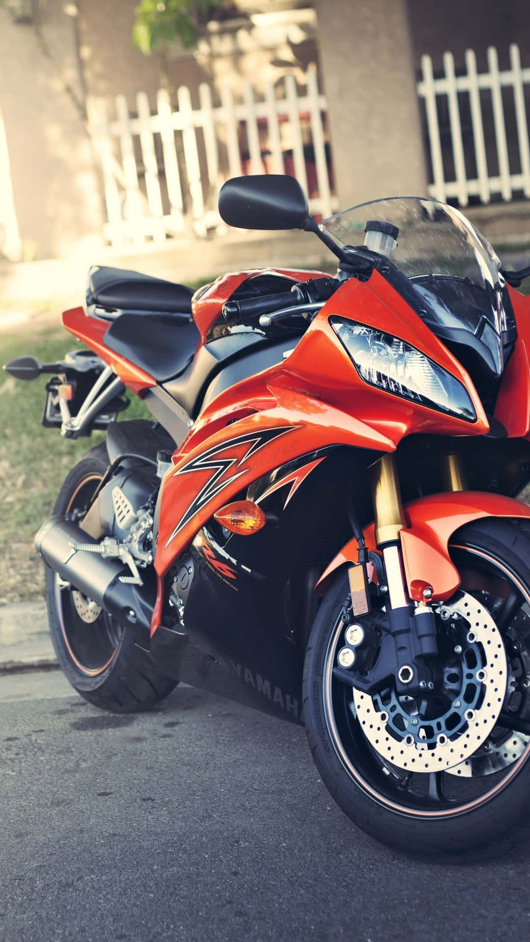  壁紙 — 一輛鮮橙色和黑色的Yamaha運動機車停放在街道上，旁邊有白色柵欄，在柔和日光下。