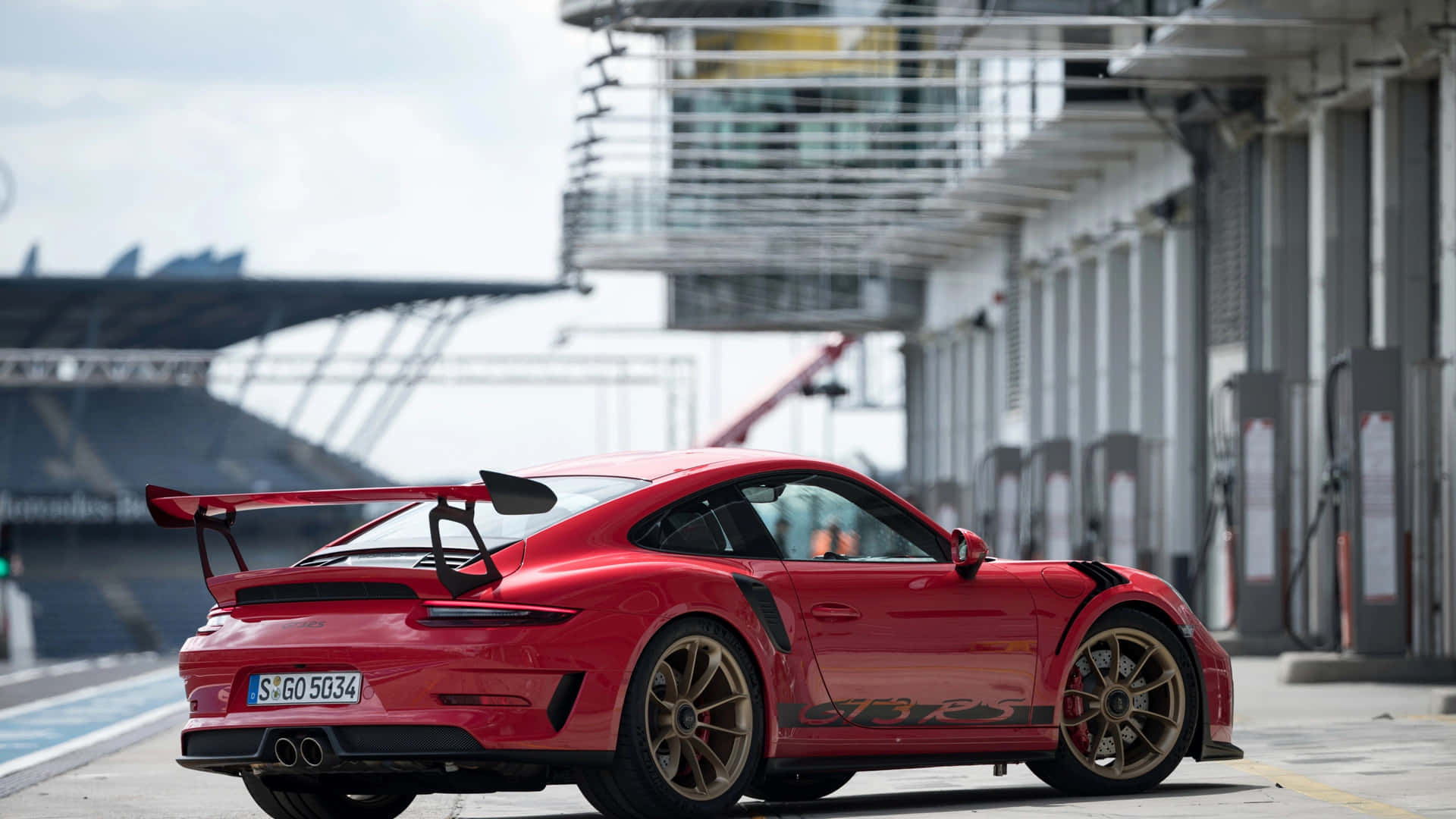  壁紙 — 一輛鮮紅色的保時捷911 GT3 RS配金色輪圈，停泊在賽車場維修區車道，天空多雲。