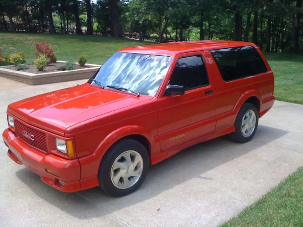 Gmc Typhoon 桌布 壁紙 — 一輛鮮紅色的GMC Typhoon運動型多功能車停在混凝土車道上，周圍環繞著綠草和樹木。