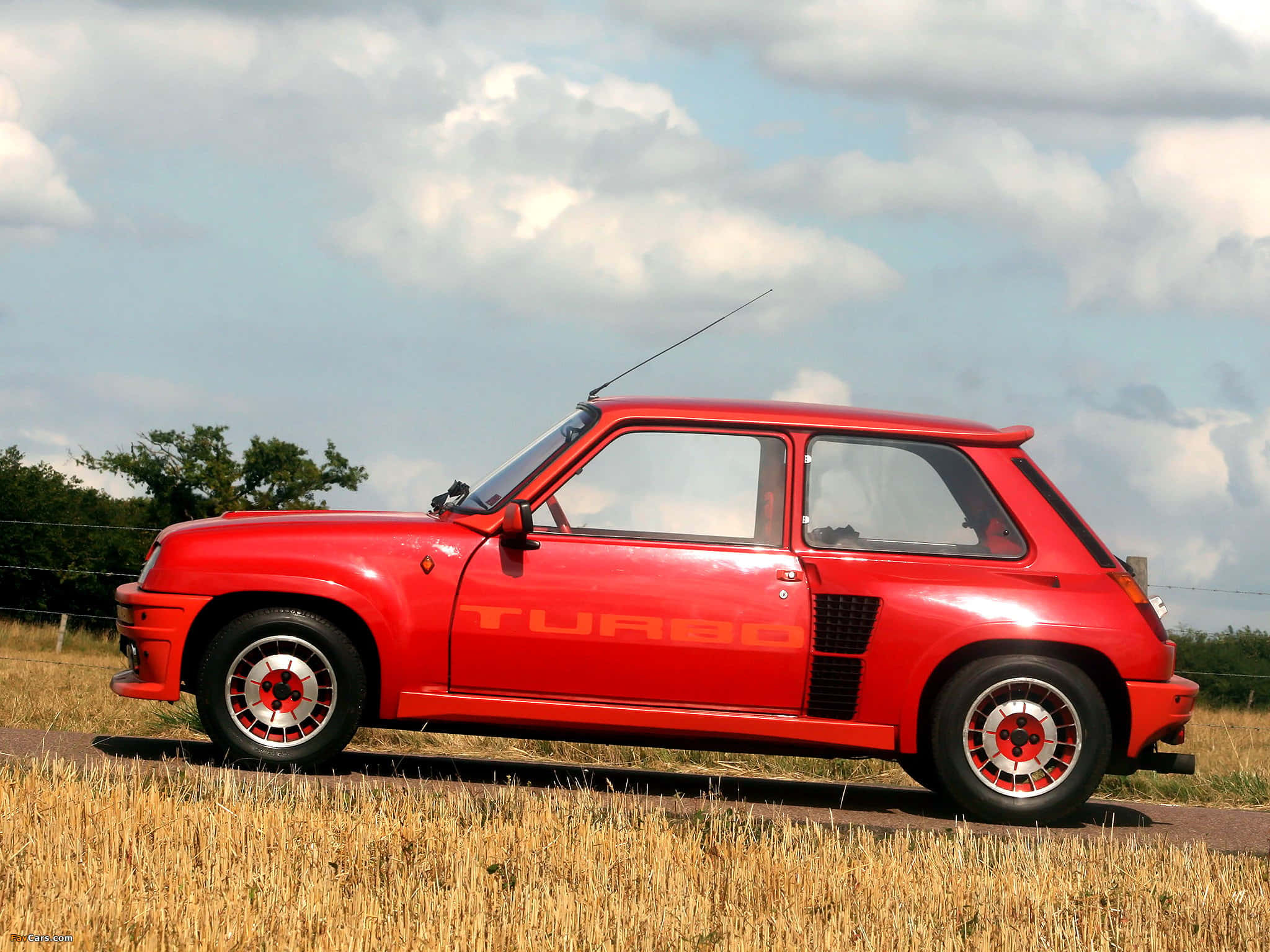 Renault 5 Turbo 桌布 壁紙 — 一輛鮮紅色的雷諾5 Turbo跑車停在鄉村道路旁，金色草叢之上，天空多雲。