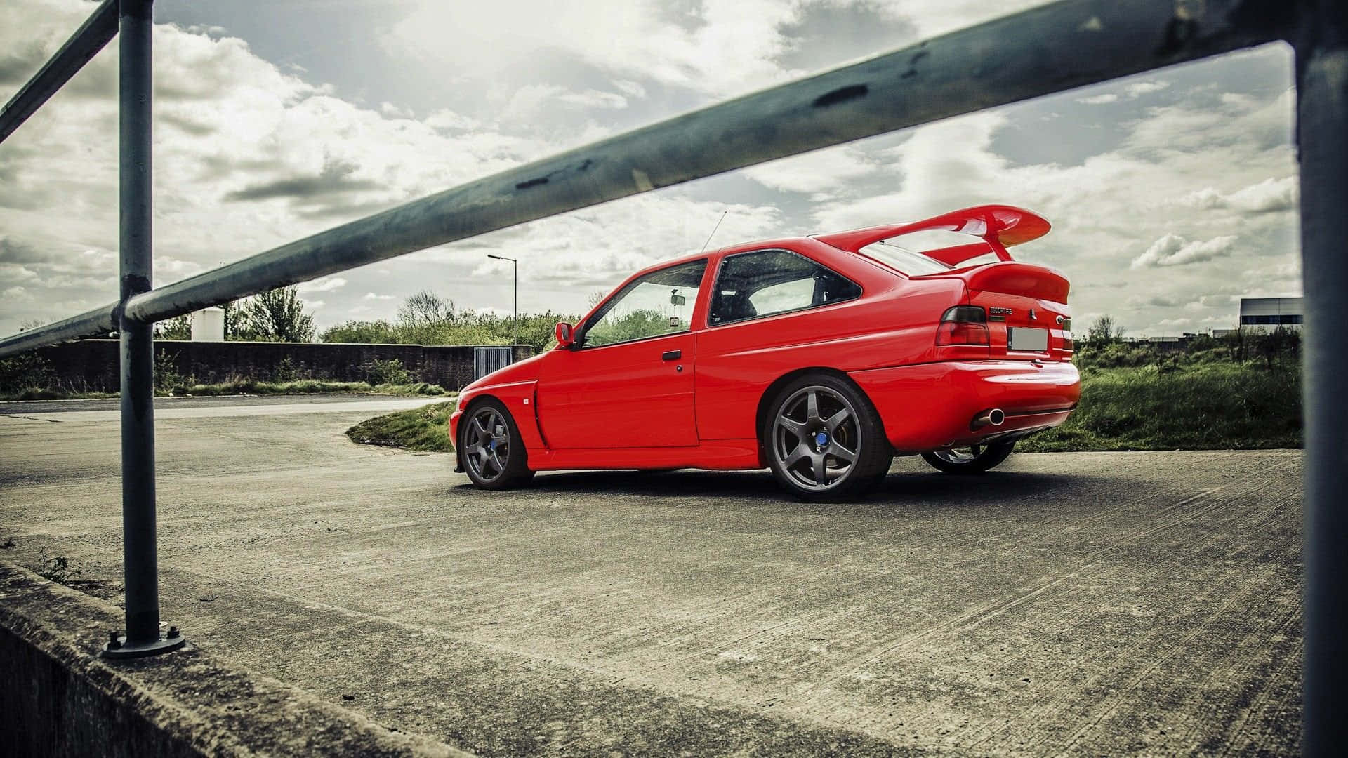 Ford Escort 桌布 壁紙 — 一輛鮮紅色的跑車，帶有大型尾翼，停泊在金屬欄杆框住的陰天之下。