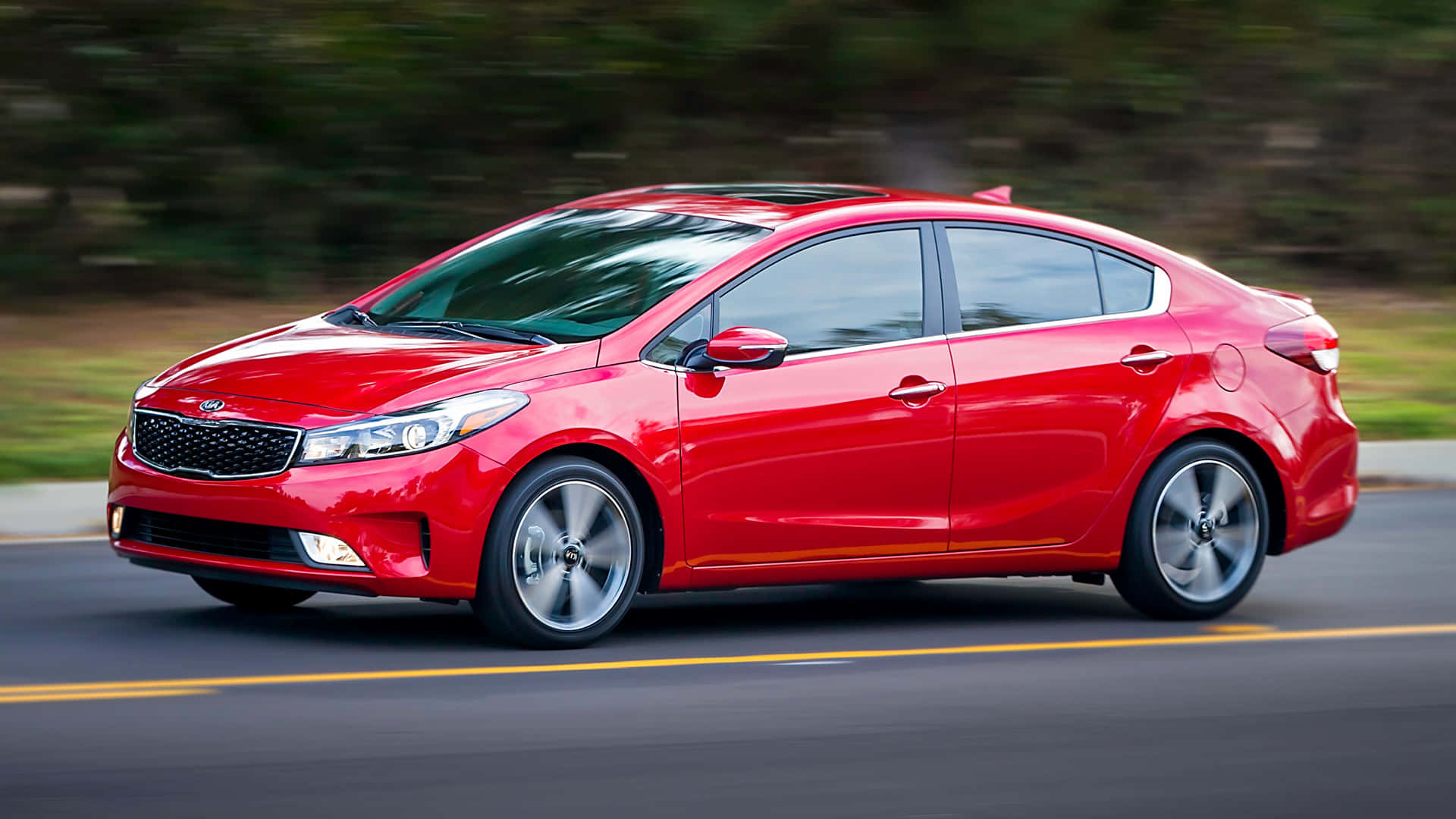 Kia Forte 桌布 壁紙 — 一輛鮮紅色的起亞轎車在柏油路上疾駛，背景是模糊的綠植。