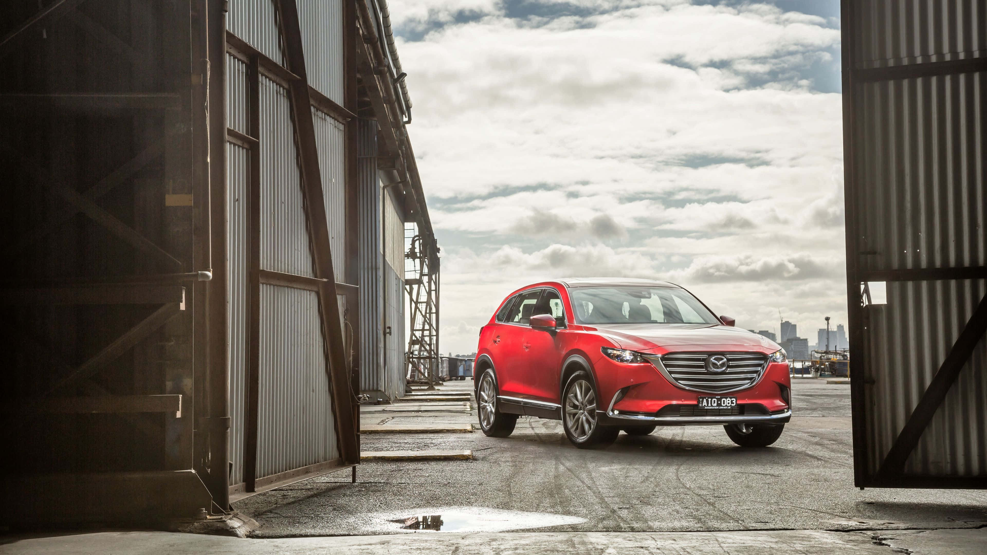 Mazda Cx-9 桌布 壁紙 — 一輛鮮紅色的Mazda CX-9 SUV停在工業金屬門之間，天空多雲，遠處有城市景觀。