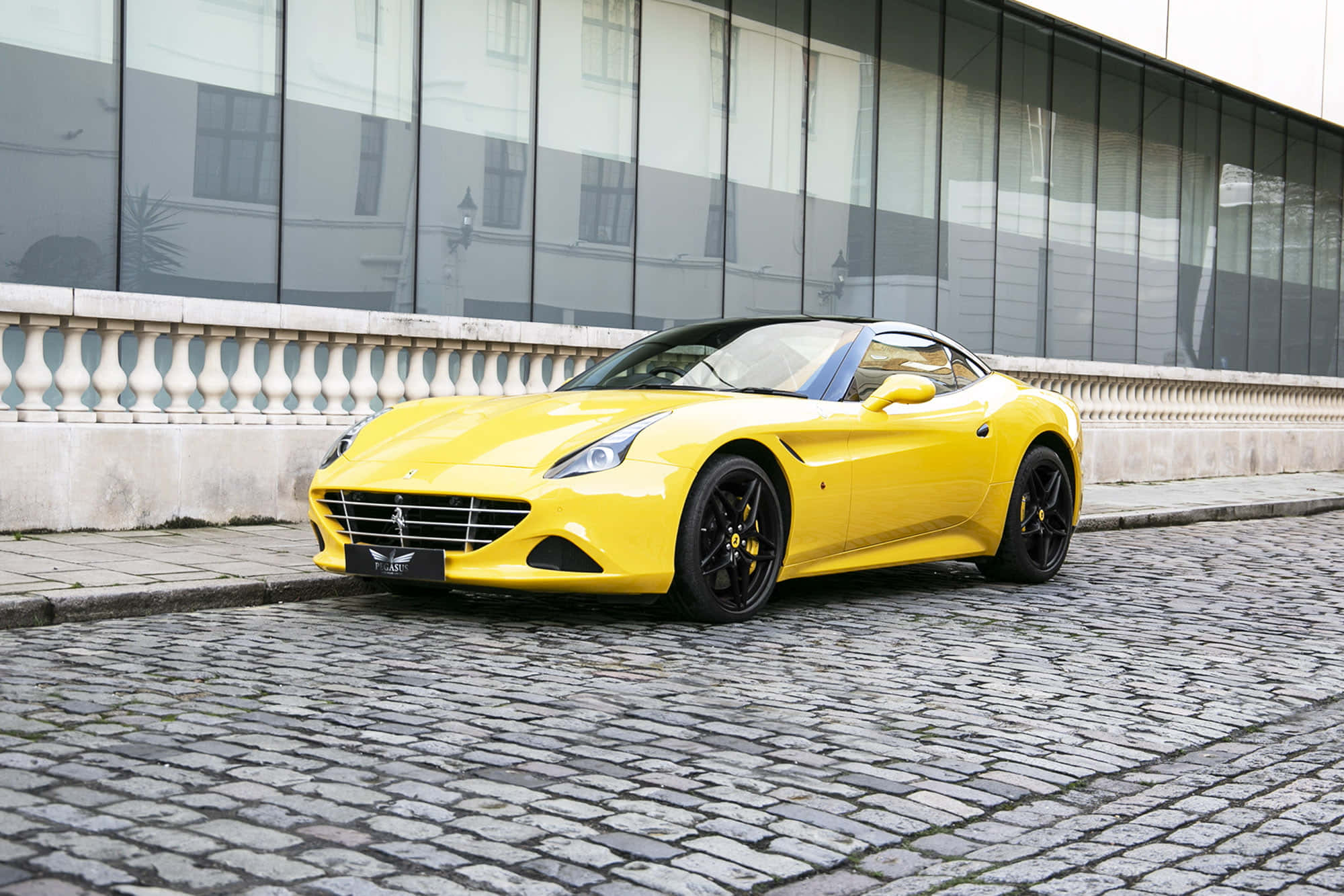 Ferrari California T 桌布 壁紙 — 一輛鮮黃色的法拉利跑車停放在現代玻璃建築旁的鵝卵石街道上。