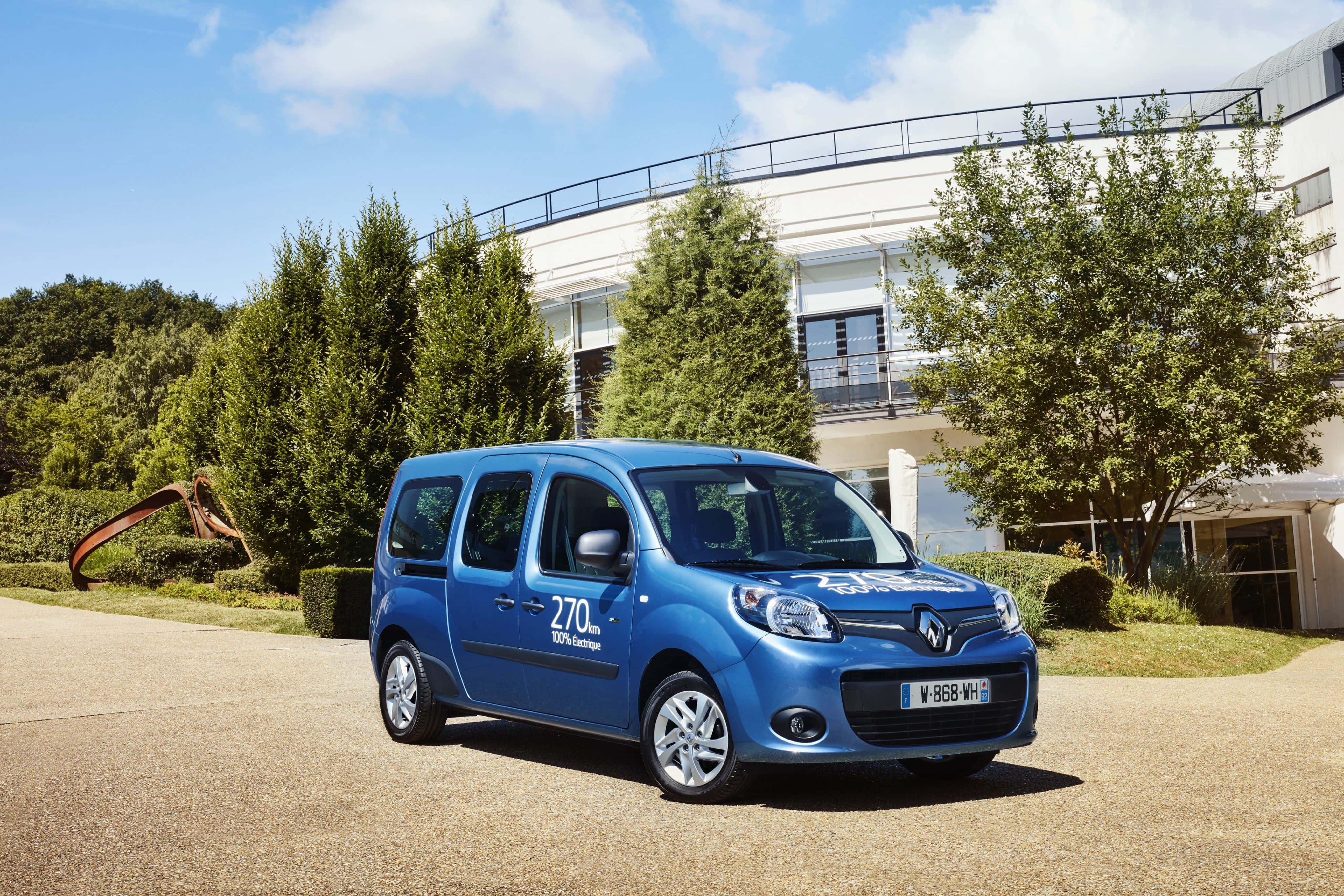 Renault Kangoo 桌布 壁紙 — 一輛鮮藍色的雷諾Kangoo停放在陽光普照的車道上，旁邊有綠樹和現代建築。