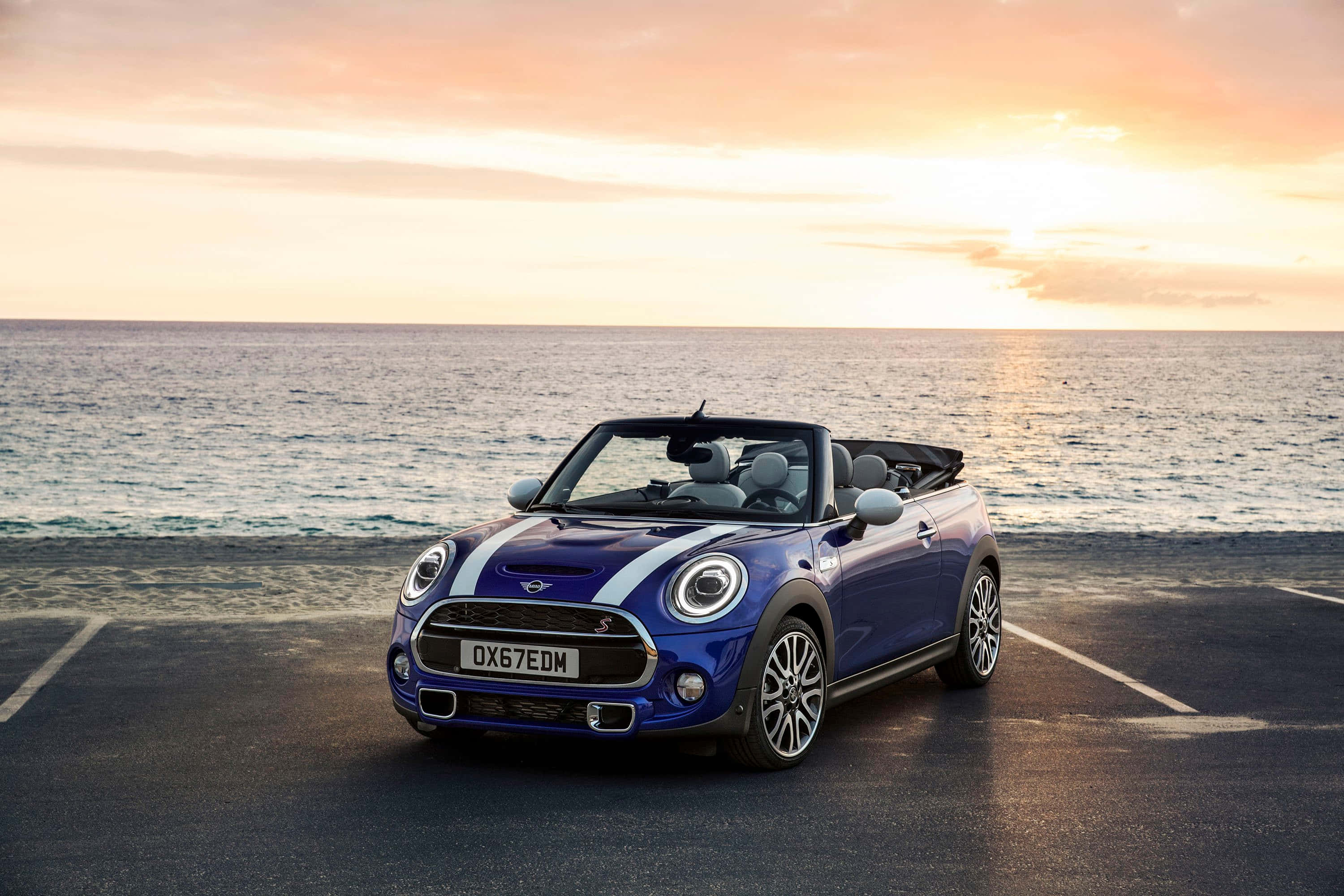 Mini Cooper Convertible 桌布 壁紙 — 一輛鮮藍色的MINI敞篷車停在日落時分的海邊道路上，海浪翻滾而來。