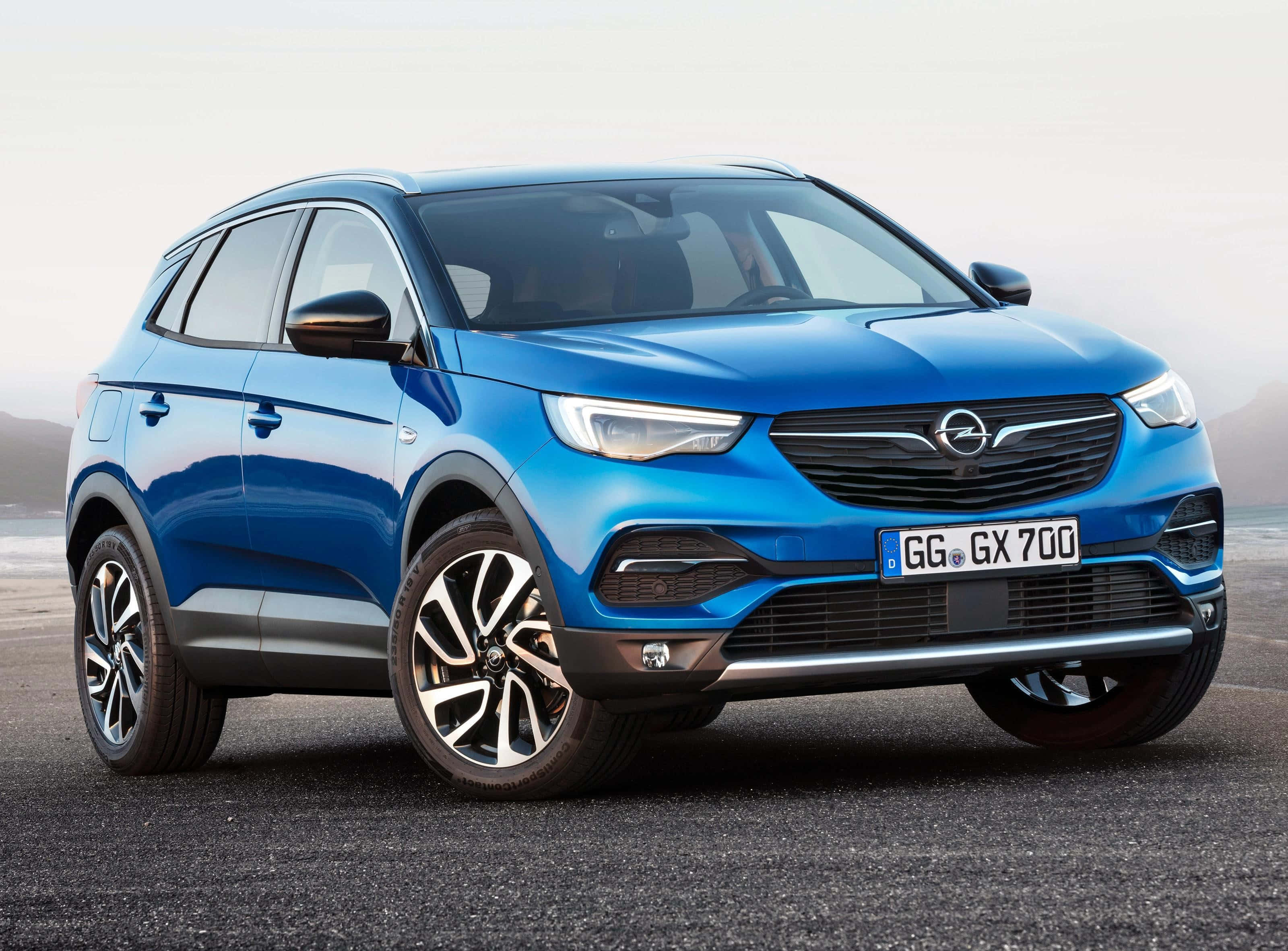 Opel Grandland X 桌布 壁紙 — 一輛鮮藍色歐寶SUV停在柏油路上，背景有朦朧的山脈，在柔和日光下。