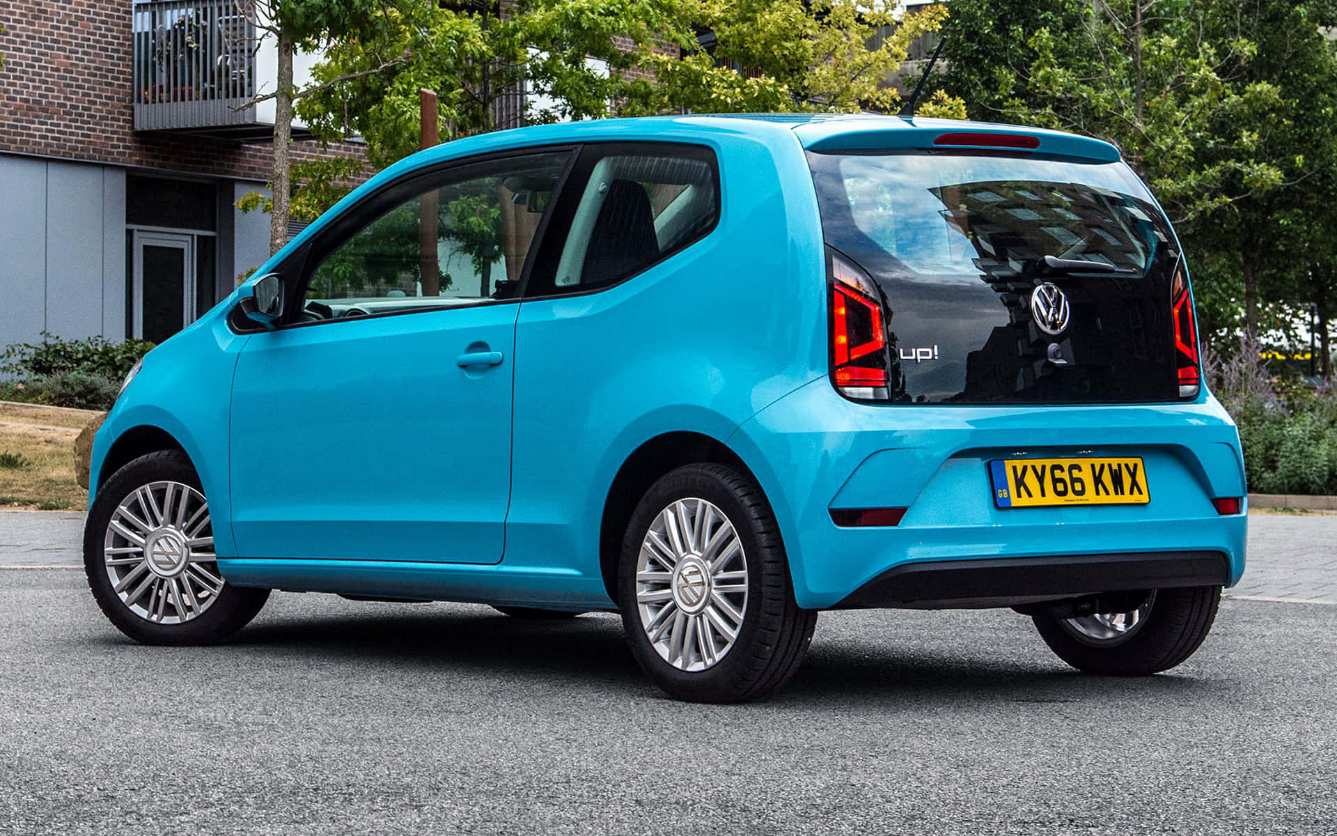  壁紙 — 一輛鮮藍色的Volkswagen Up! 停在柏油路上，背景有樹木和磚造建築。