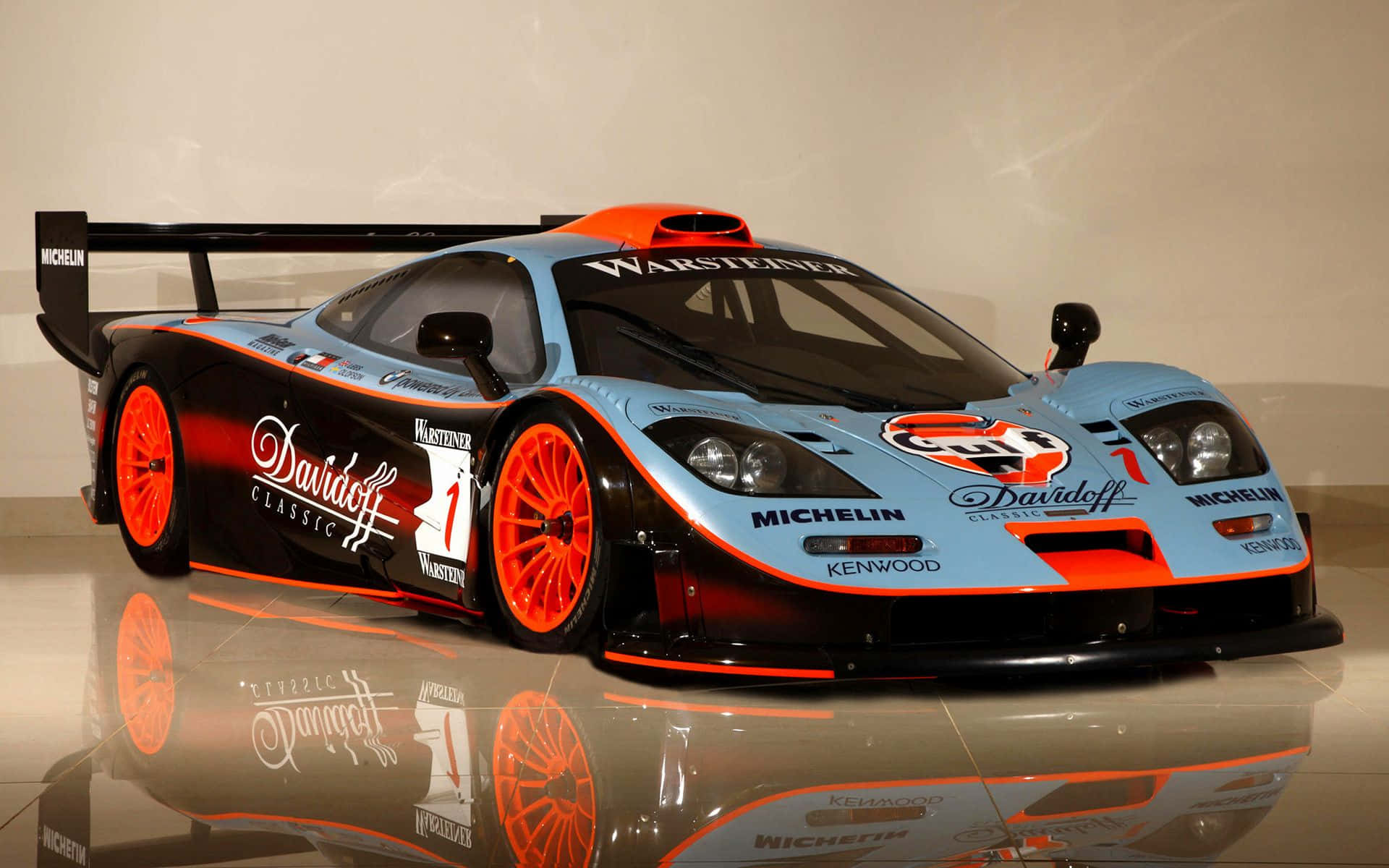 Mclaren F1 桌布 壁紙 — 一輛線條流暢的McLaren賽車，配備橙色輪圈和藍黑塗裝，在博物館的反射地板上展示。