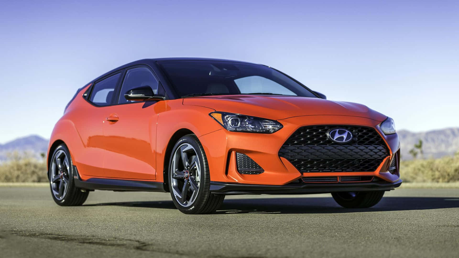 Hyundai Veloster 桌布 壁紙 — 一輛鮮豔的橙色現代Veloster N跑車停在瀝青路上，背景是山脈，天空清澈湛藍。