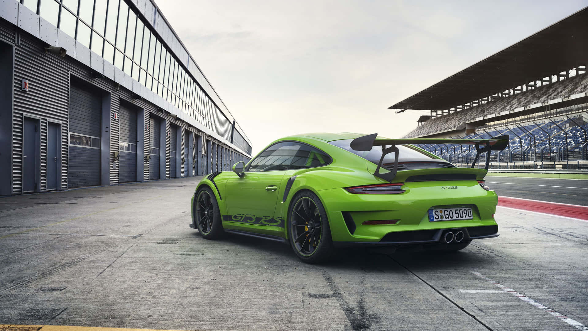  壁紙 — 一輛鮮豔的青檸綠色保時捷911 GT3 RS停泊在賽道上，旁邊有看台和車庫建築，天空多雲。