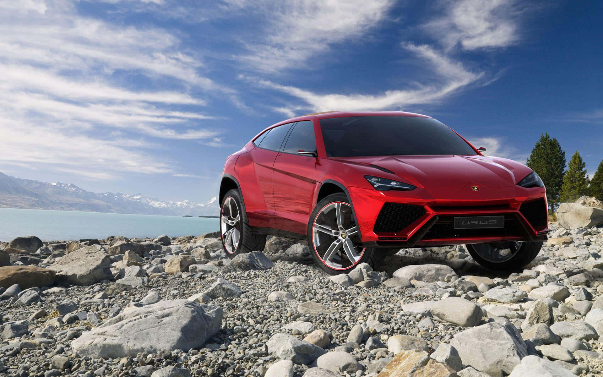 Lamborghini Urus 桌布 壁紙 — 一輛醒目的紅色藍寶堅尼 Urus SUV 大膽地停在岩石湖岸上，背景是白雪覆蓋的山脈和明亮的藍天。