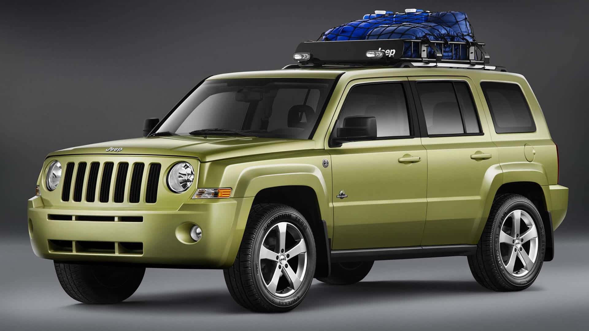 Jeep Patriot 桌布 壁紙 — 一輛啞光橄欖綠色的Jeep Patriot SUV，車頂架上有一個藍色貨物箱，站在深灰色工作室背景前。