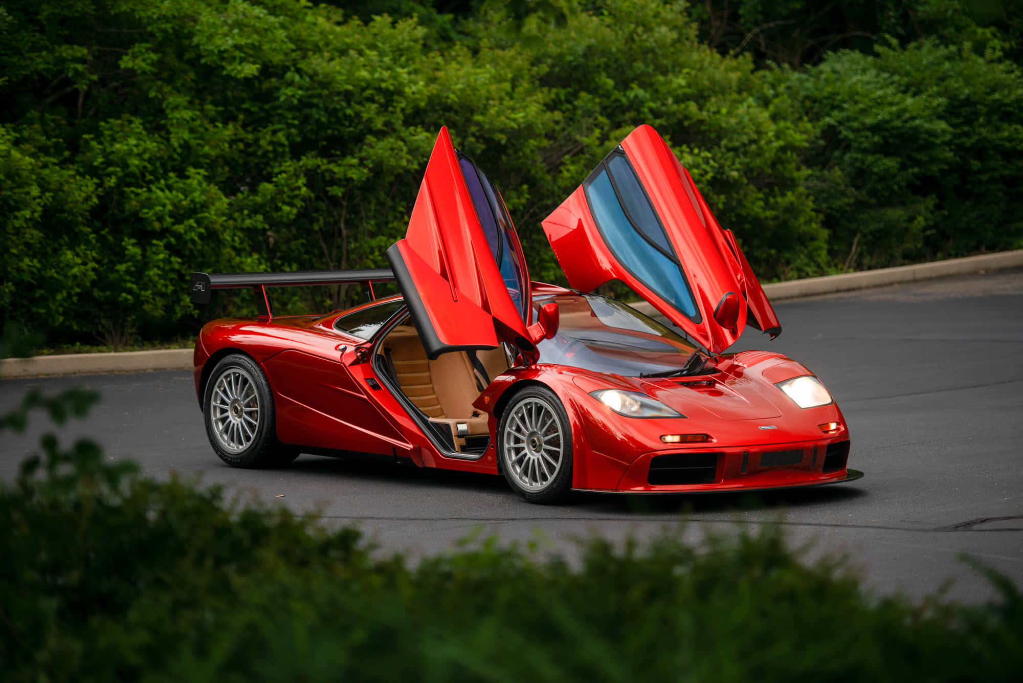 Mclaren F1 桌布 壁紙 — 一輛引人注目的紅色McLaren F1超級跑車，鷗翼車門打開，停泊在綠樹成蔭的道路上。