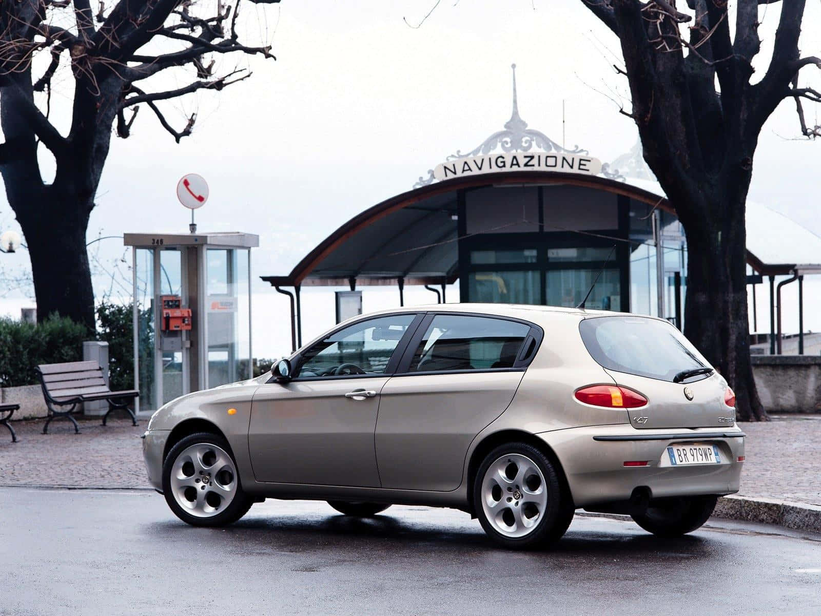 Alfa Romeo 147 桌布 壁紙 — 一輛銀色愛快羅密歐147停泊在濕漉漉的路面上，旁邊有電話亭和一個「Navigazione」候車亭，天空多雲。