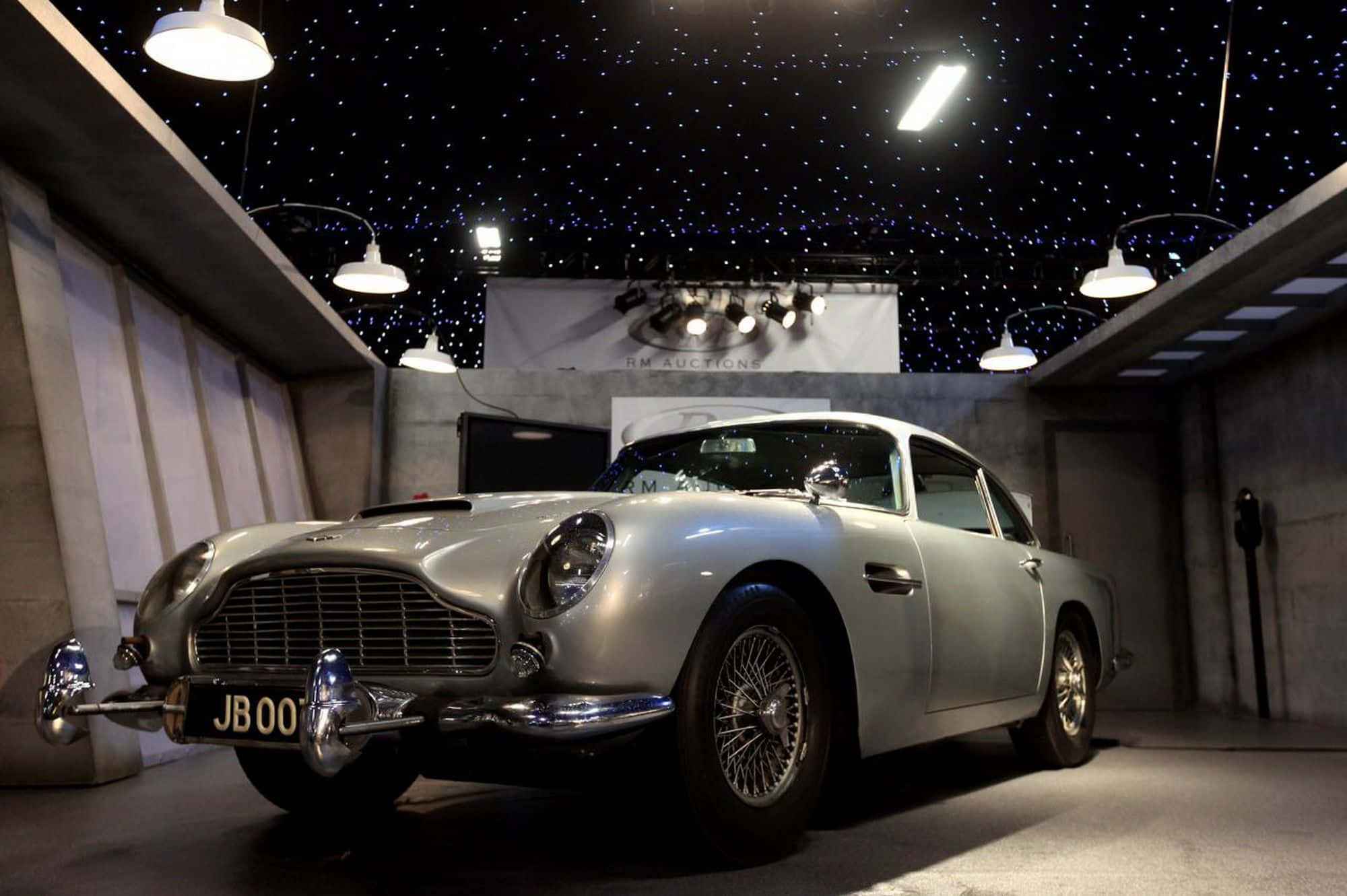 Aston Martin Db5 桌布 壁紙 — 一輛銀色Aston Martin DB5在混凝土拍賣畫廊的星空天花板下，在聚光燈下閃閃發光。