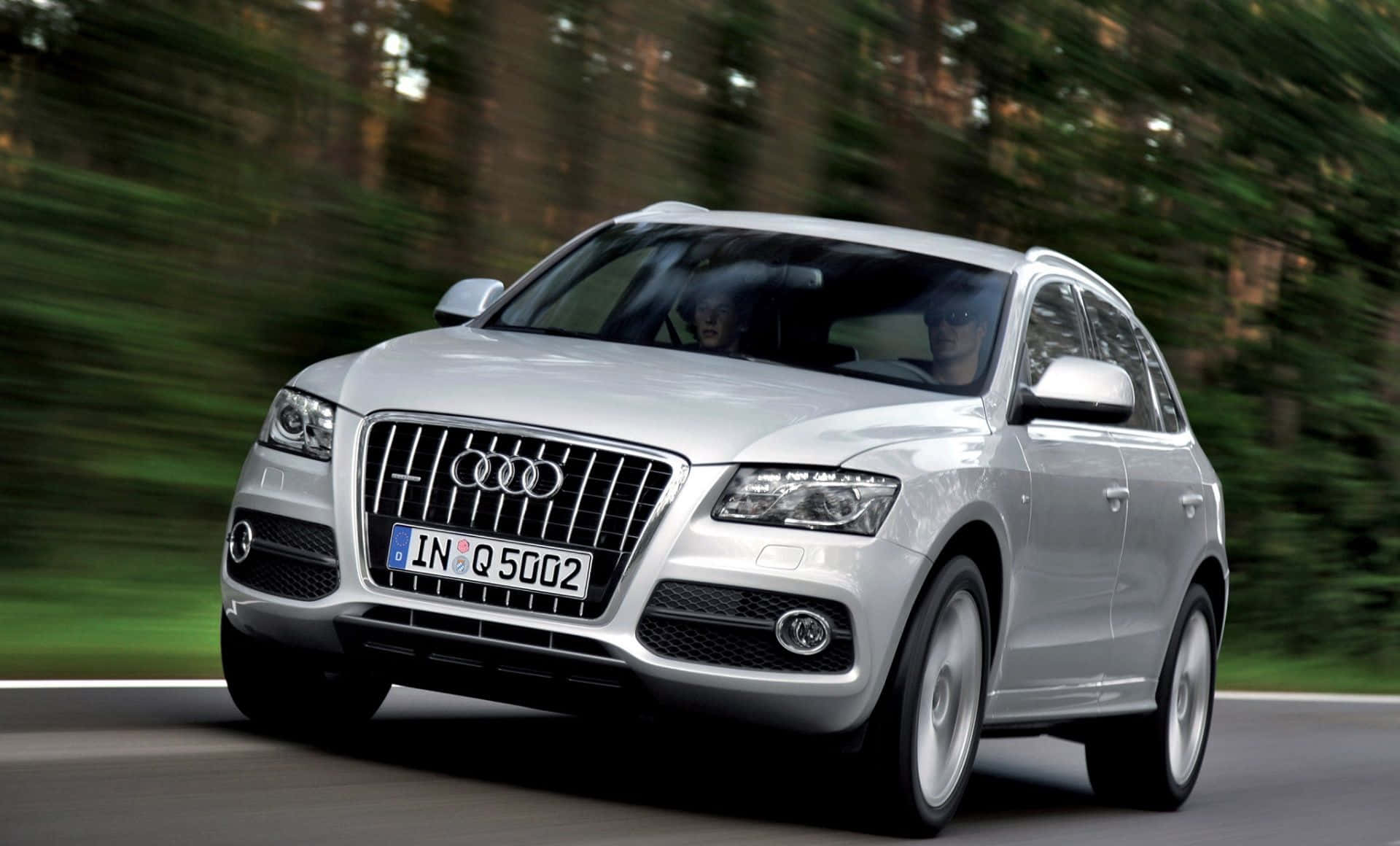Audi Q5 桌布 壁紙 — 一輛銀色 Audi Q5 SUV 在模糊的綠色森林背景中沿著道路高速行駛。