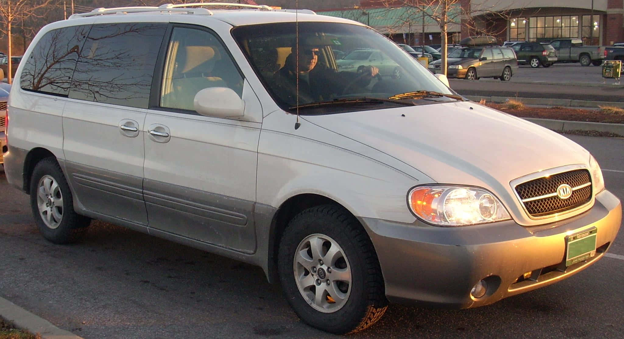 Kia Sedona 桌布 壁紙 — 一輛銀色Kia小型貨車停放在一個停車場，背景有光禿禿的樹木和其他車輛。