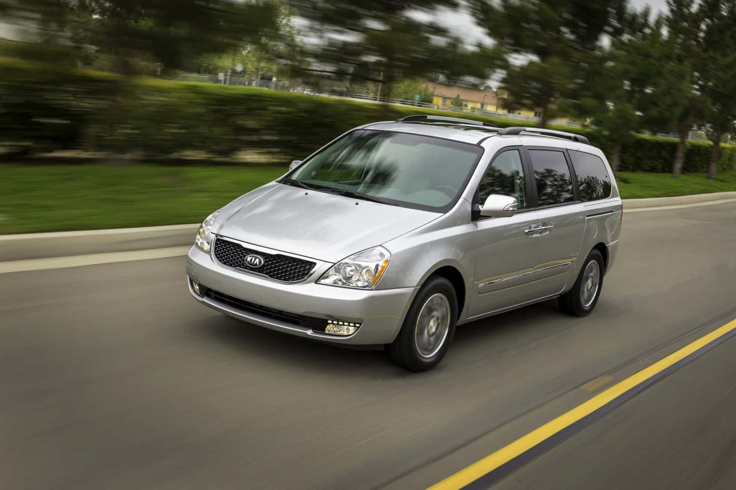 Kia Sedona 桌布 壁紙 — 一輛銀色Kia小型貨車在郊區道路上疾駛，背景有模糊的綠化和房屋。