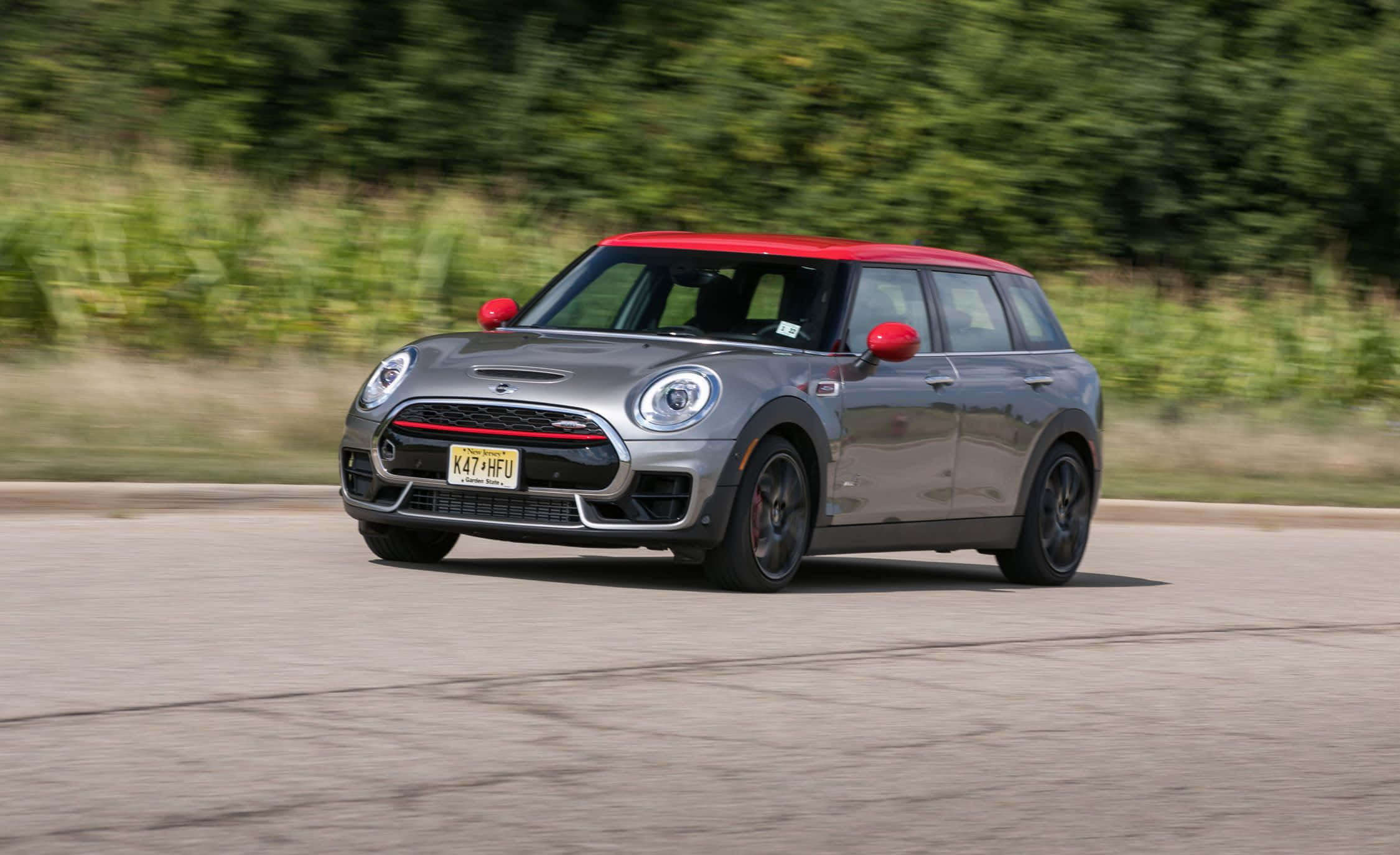 Mini Cooper S Clubman All4 桌布 壁紙 — 一輛銀色MINI Cooper，紅色車頂和後視鏡，在柏油路上奔馳，背景是模糊的綠色樹葉。