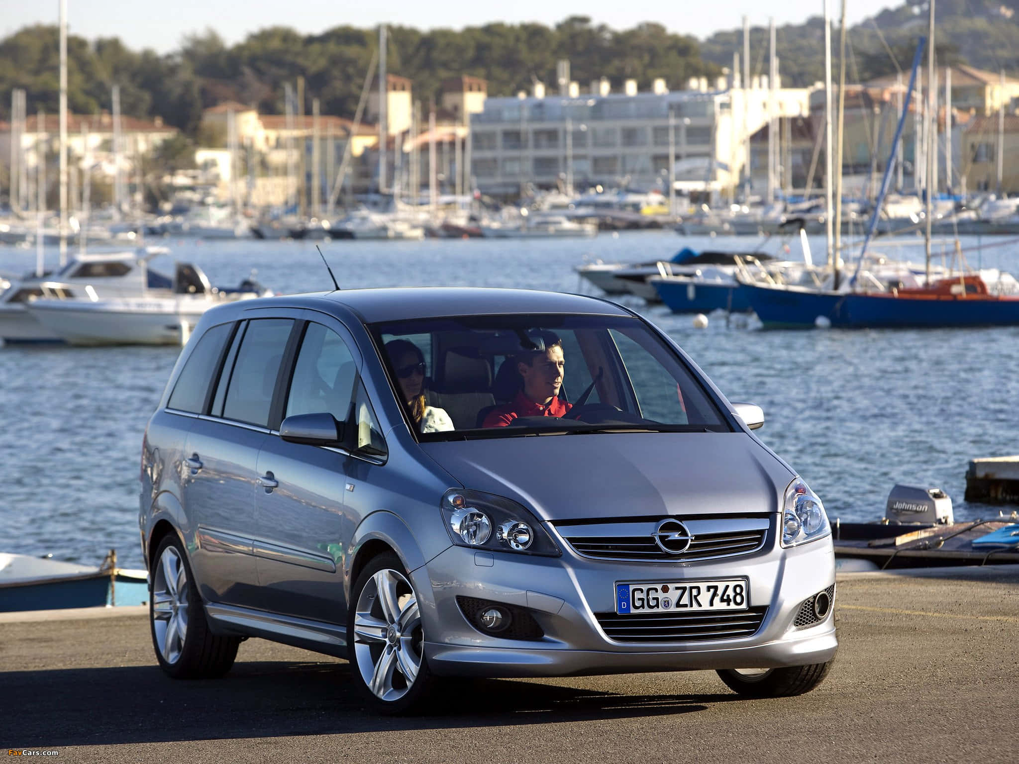 Opel Zafira 桌布 壁紙 — 一輛銀色歐寶Zafira在明亮的日光下行駛在海濱道路上，背景有帆船和建築物。