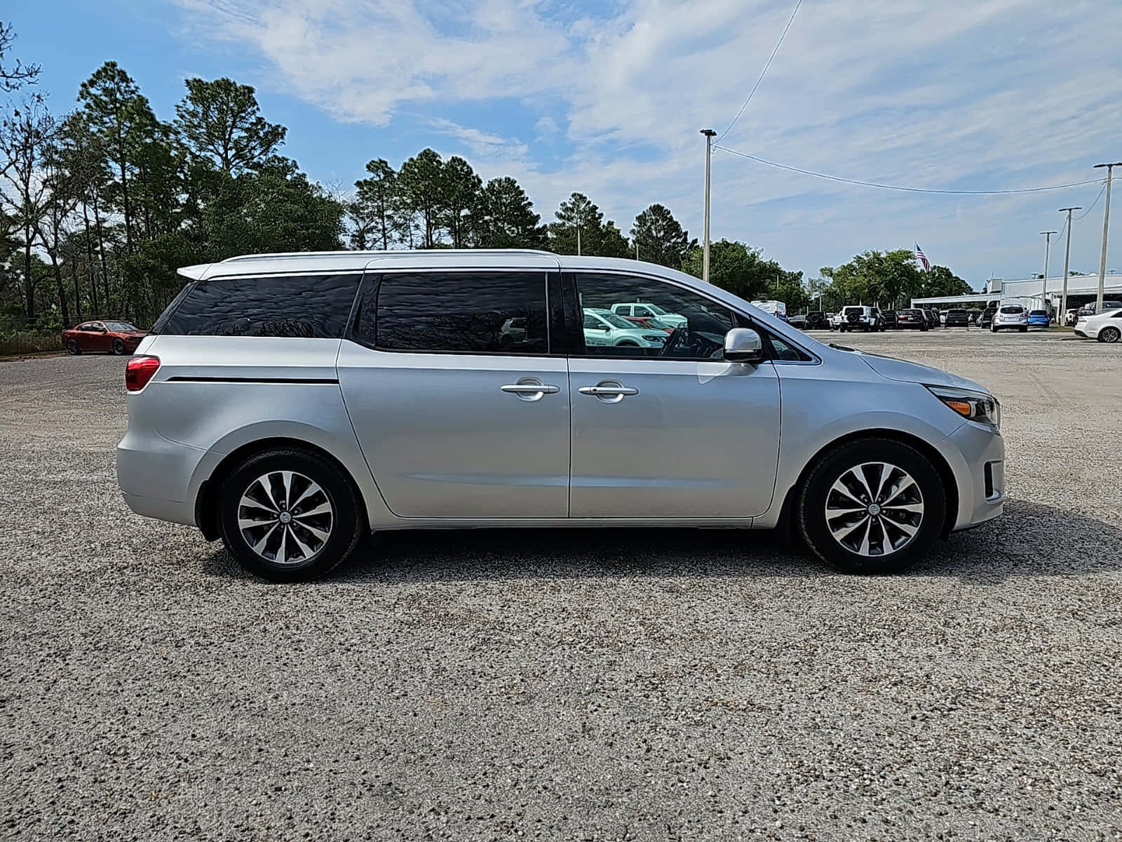 Kia Sedona 桌布 壁紙 — 一輛銀色小型貨車停在碎石上，背景有樹木和部分多雲的天空。