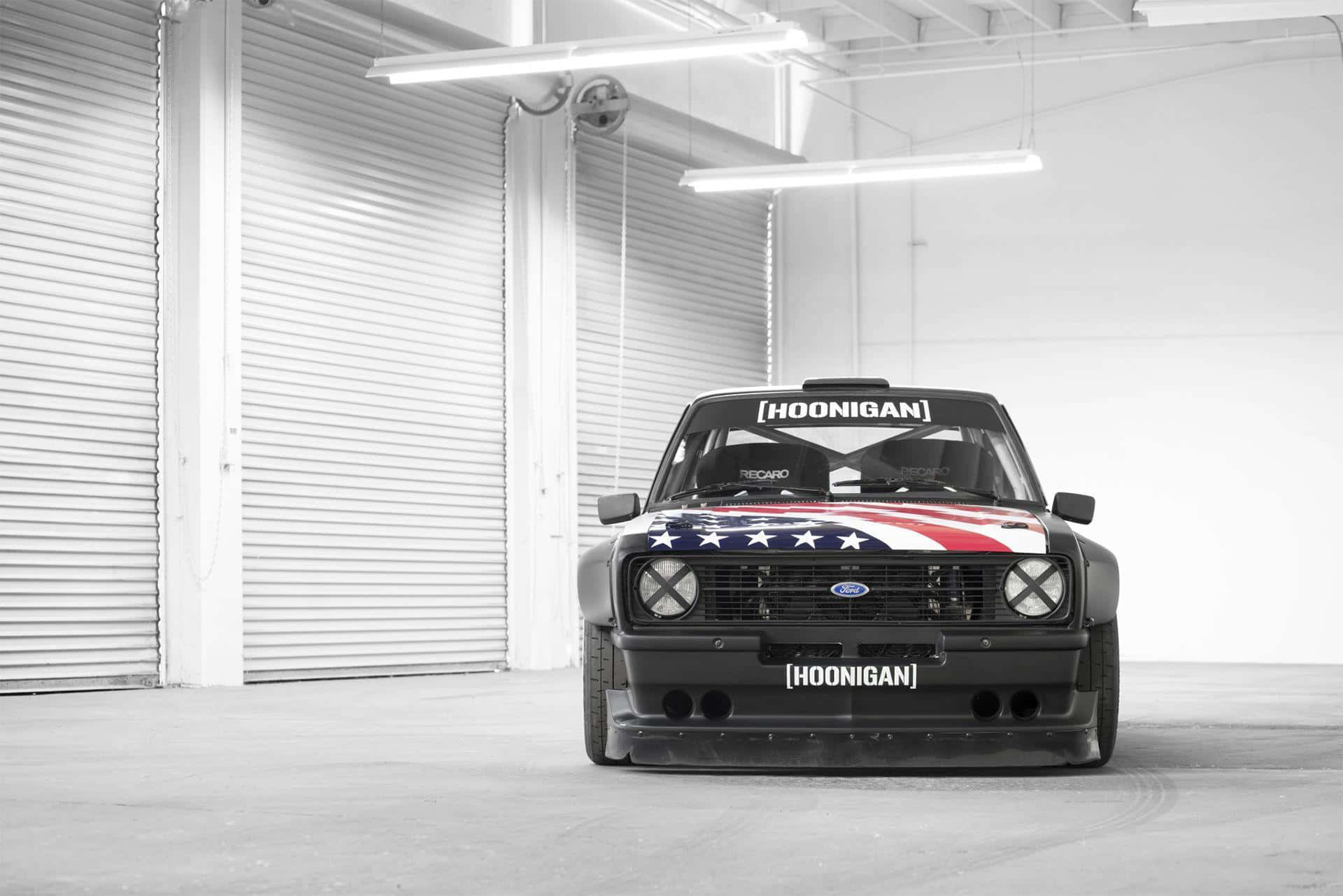 Ken Block 桌布 壁紙 — 一輛帶有美國國旗塗裝的黑色Hoonigan Ford拉力賽車，在螢光燈下停放在明亮的工業車庫中央。