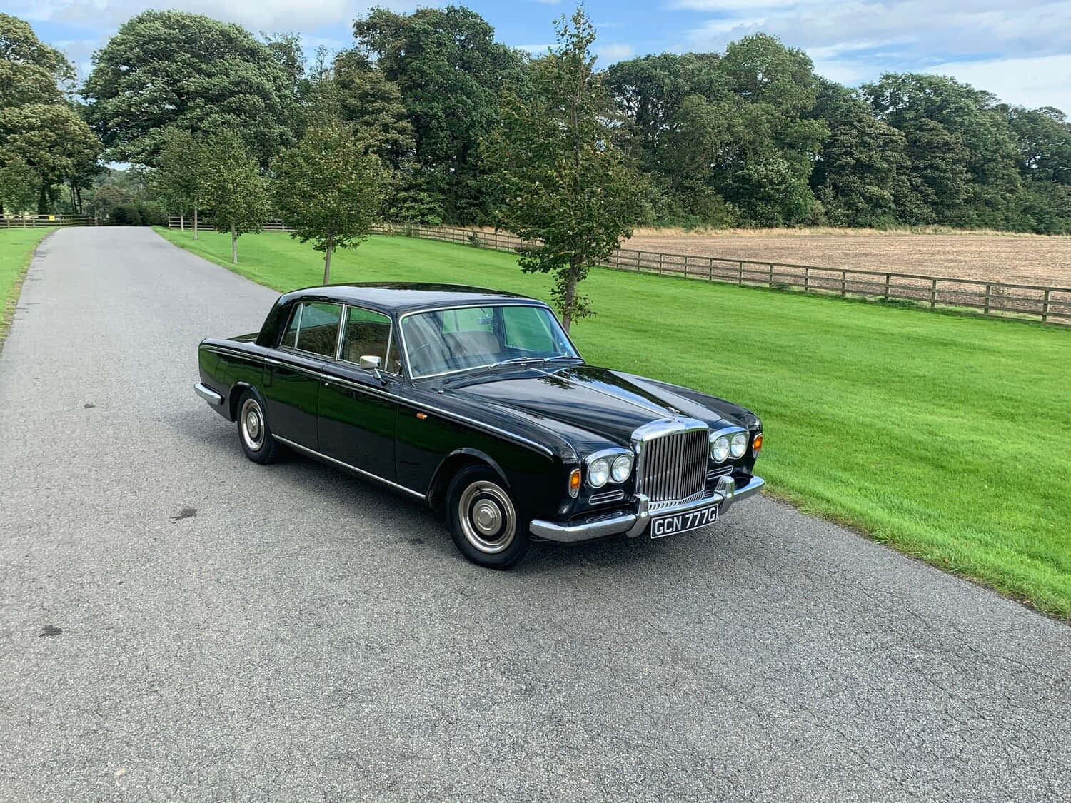 Bentley T1 桌布 壁紙 — 一輛光澤黑色經典賓利轎車停在碎石路上，旁邊是修剪整齊的綠色草坪和枝葉茂密的樹木，天空部分多雲。