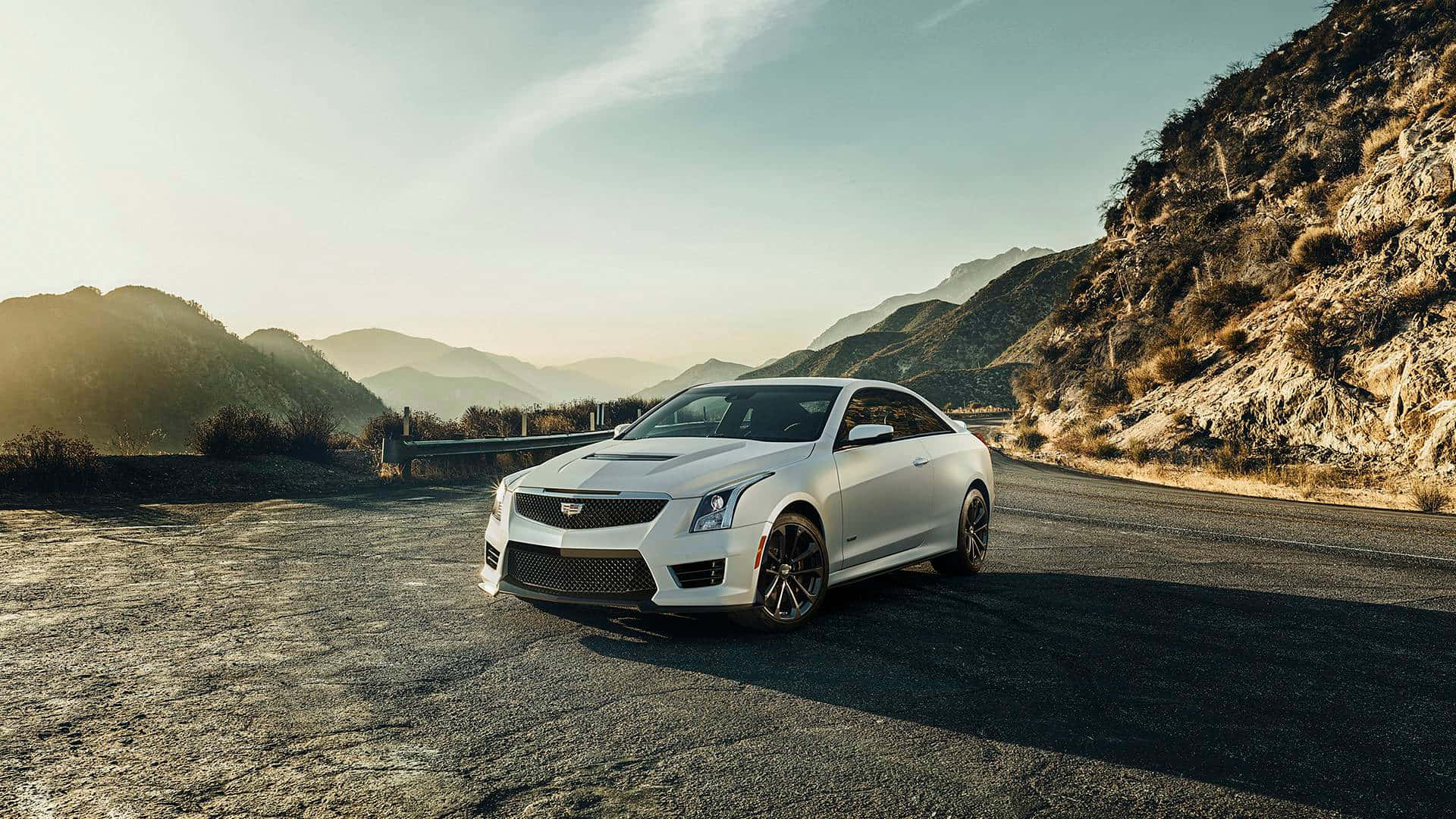 Cadillac Xts 桌布 壁紙 — 一輛時尚的白色Cadillac ATS-V雙門跑車停泊在蜿蜒的山路上，正值黃金時刻，崎嶇的懸崖和遠處的山峰沐浴在溫暖的陽光中。