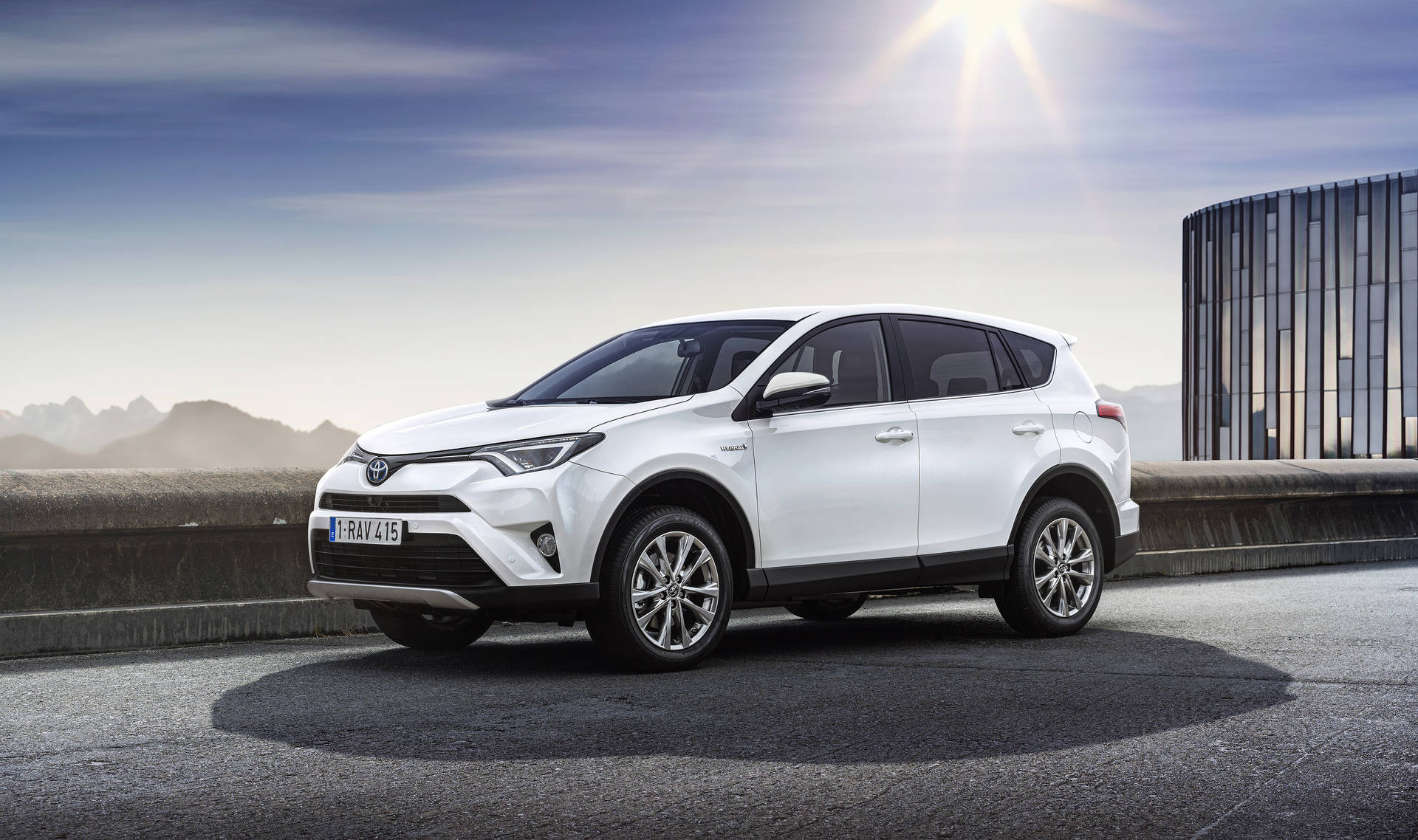 白色豐田RAV4 SUV在橋上，背景有山脈和現代建築。 — Toyota Supra 壁紙