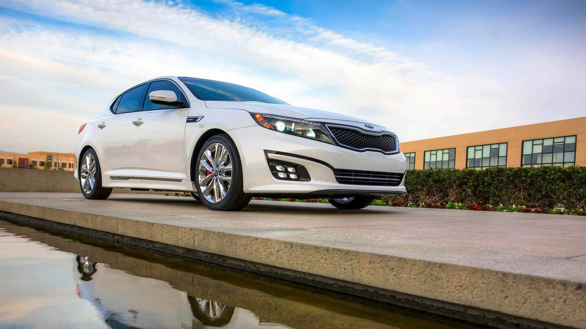 Kia Optima 桌布 壁紙 — 一輛時尚的白色Kia Optima轎車在藍天白雲下閃閃發光，停泊在一個反射水景旁邊。