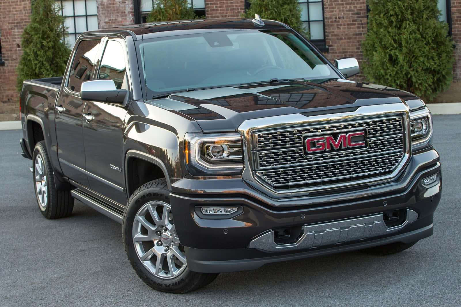 Gmc Sierra 桌布 壁紙 — 一輛時尚的黑色GMC Sierra皮卡在日光下閃耀，停在有綠色灌木的磚砌建築前。