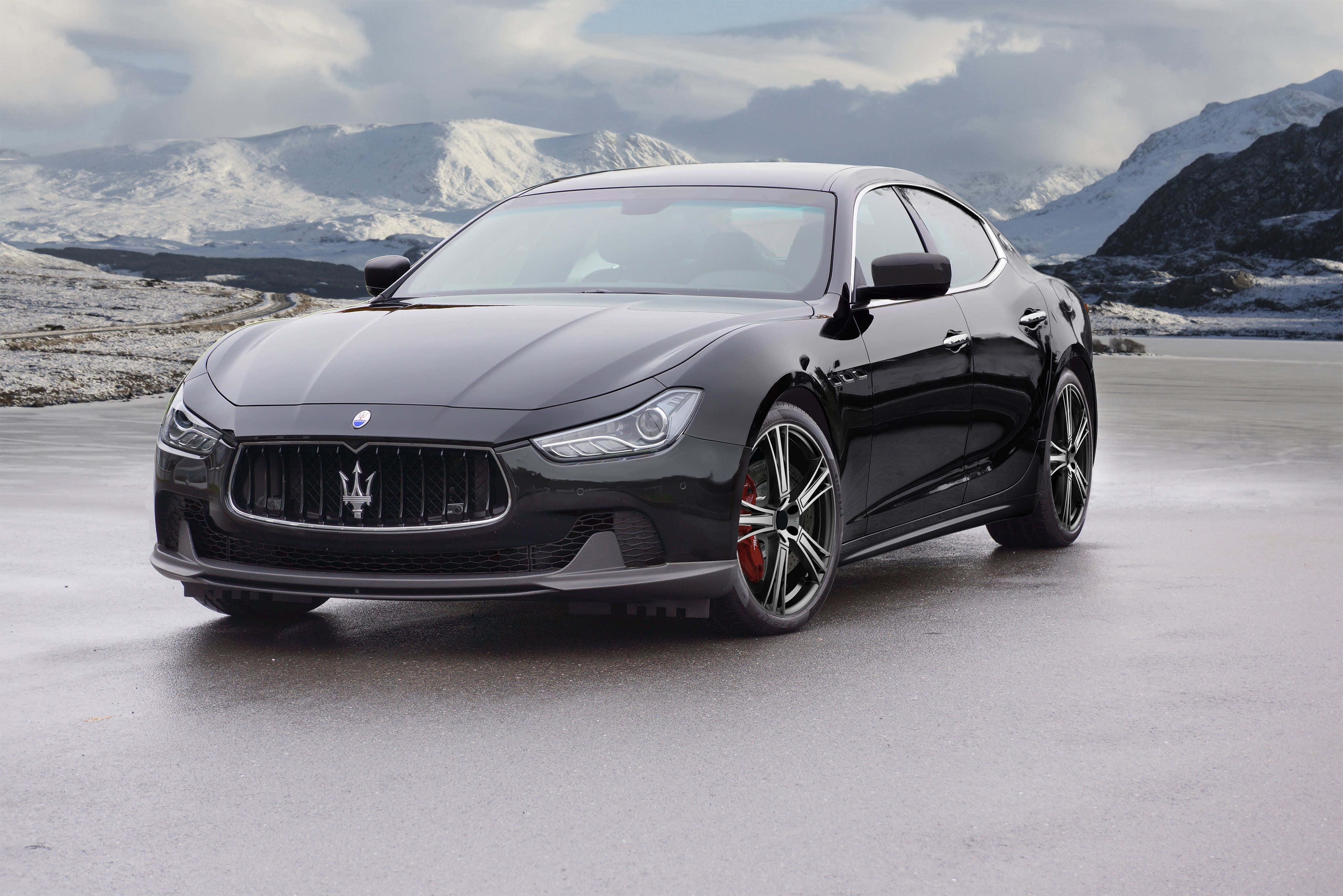 Maserati Ghibli 桌布 壁紙 — 一輛時尚的黑色瑪莎拉蒂跑車在潮濕的道路上閃閃發光，白雪覆蓋的山脈在陰雲密布的天空下若隱若現。