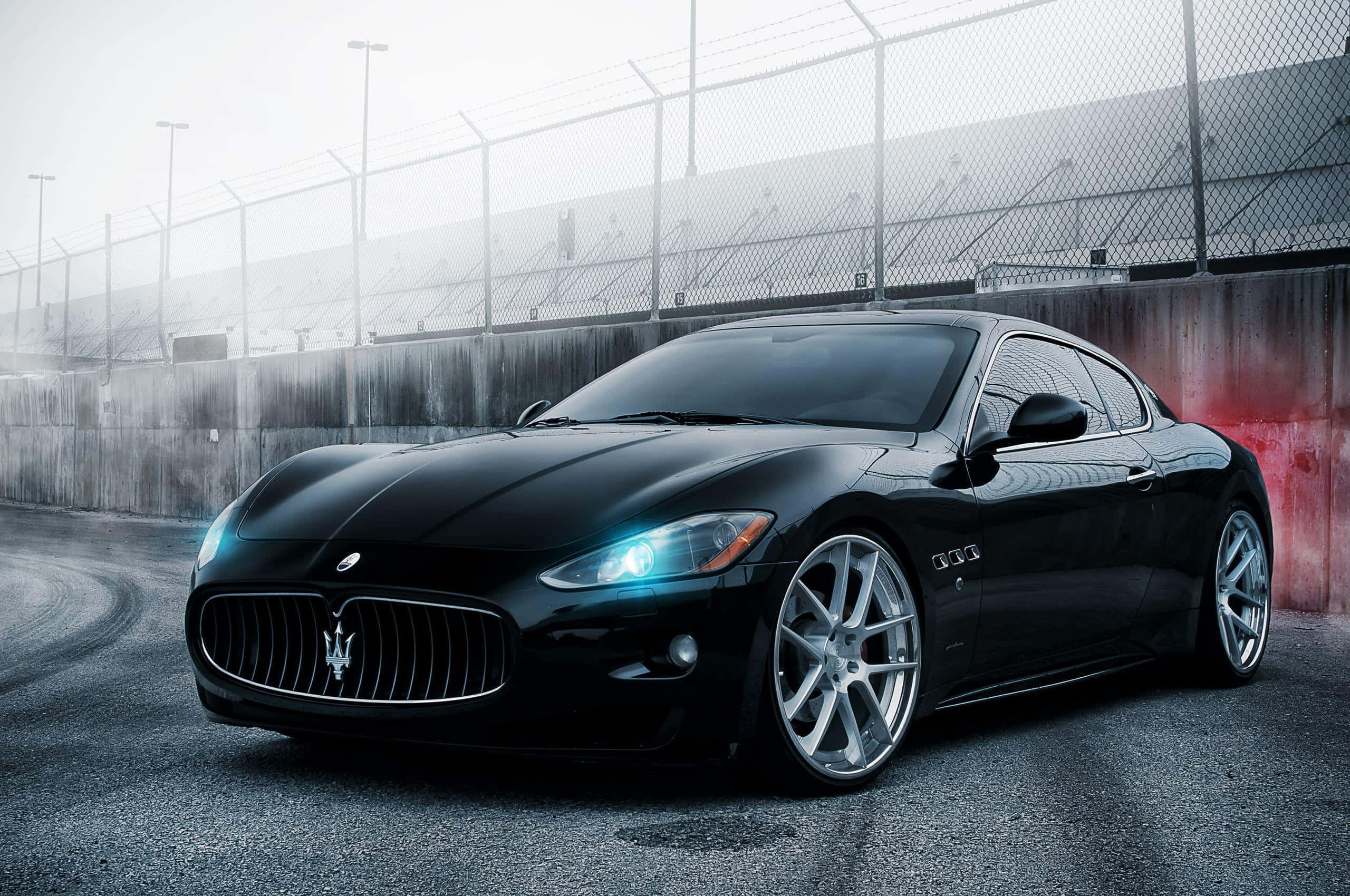 Maserati Quattroporte 桌布 壁紙 — 一輛時尚的黑色Maserati跑車，車頭燈閃爍，停泊在濕滑的賽道上，旁邊是混凝土牆和鐵絲網圍欄。