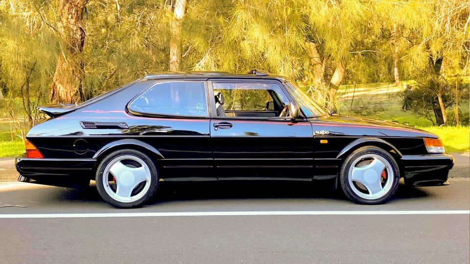 Saab 99 桌布 壁紙 — 一輛時尚的黑色Saab 900 Turbo停泊在陽光普照的道路旁，旁邊是金黃色的樹木。