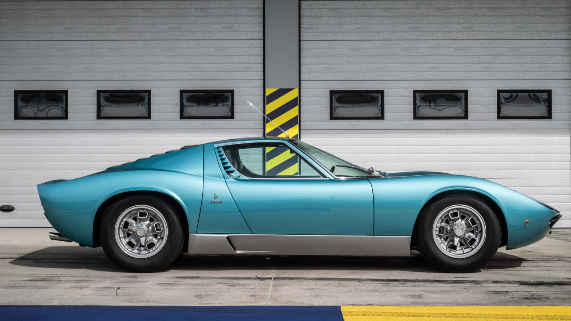 Lamborghini Miura 桌布 壁紙 — 一輛時尚的藍綠色經典跑車停放在灰色工業車庫門前，門上有黃色安全條紋。