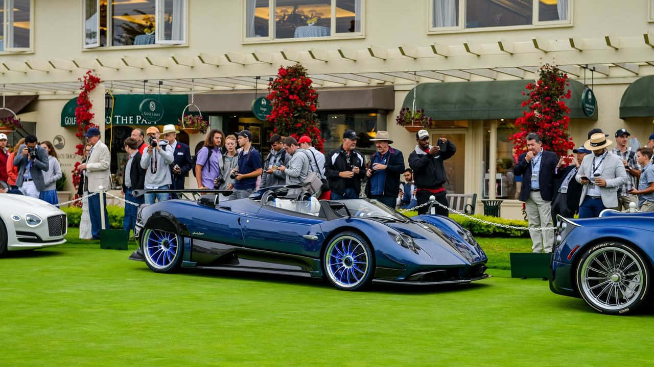 Pagani Zonda Hp Barchetta 桌布 壁紙 — 一輛時尚的藍色Pagani Huayra敞篷跑車在修剪整齊的綠色草地上閃閃發光，攝影師和觀眾聚集在豪華車展活動中。