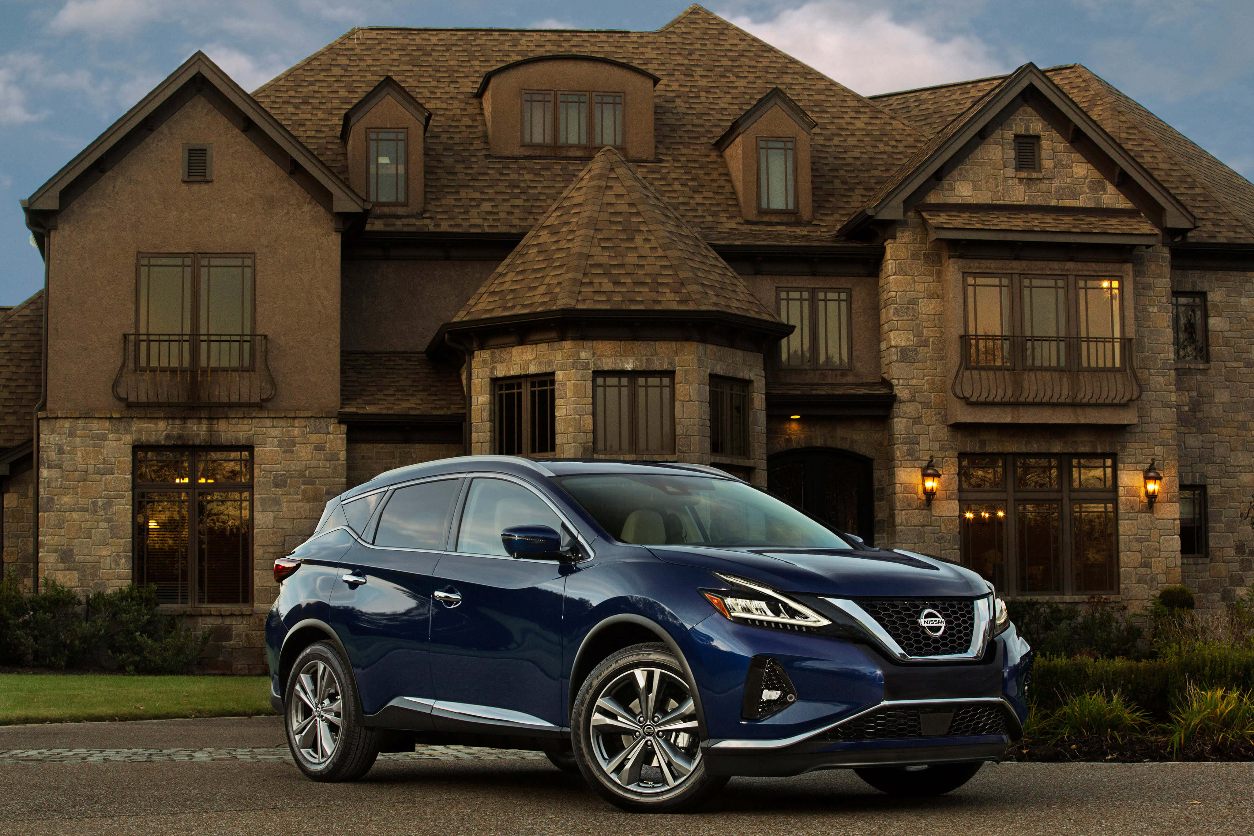 Nissan Murano 桌布 壁紙 — 一輛時尚的藍色日產Rogue SUV停在一棟大型石砌豪宅前，溫暖的晚燈在窗戶中閃爍。