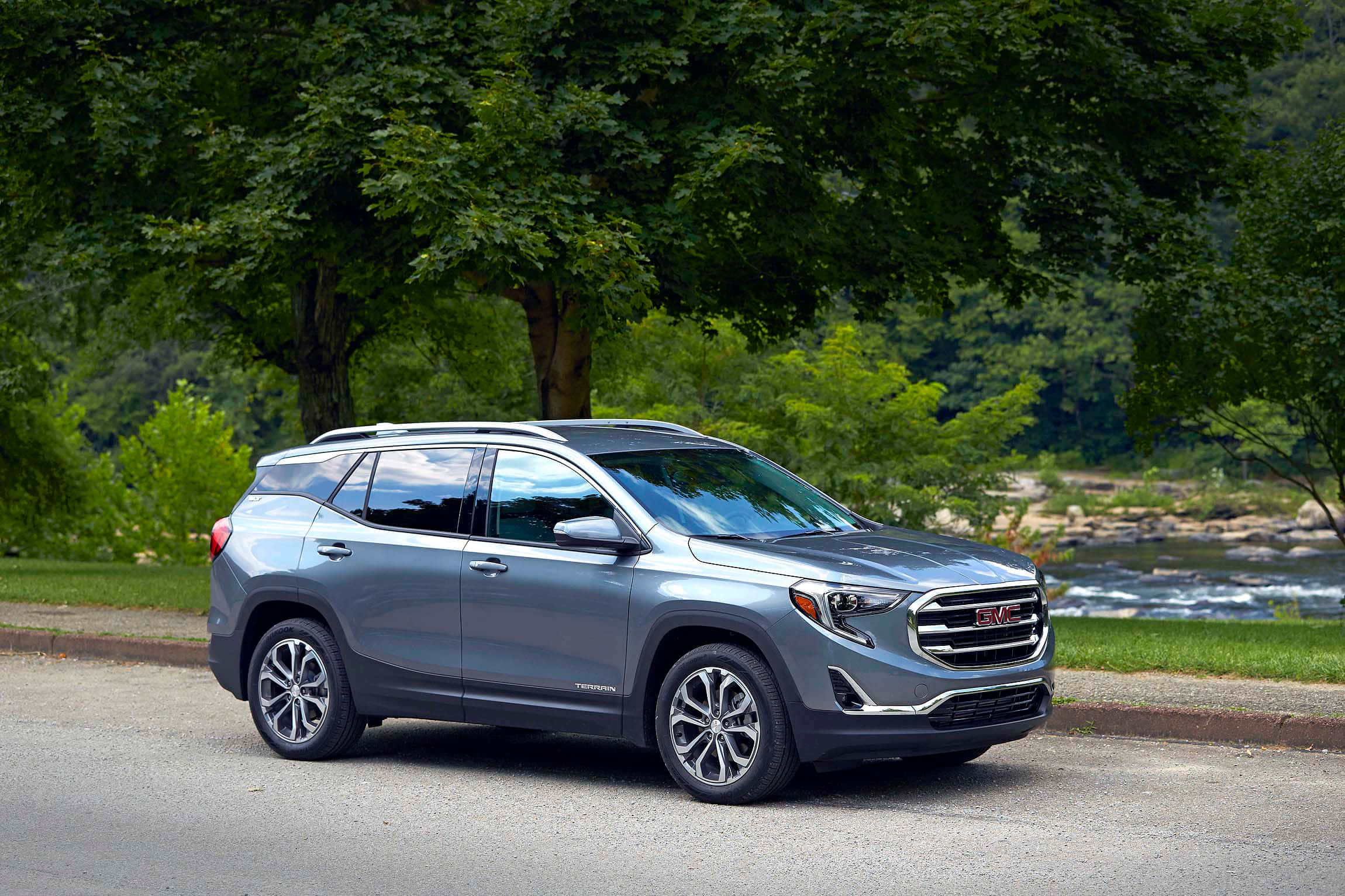 Gmc Terrain 桌布 壁紙 — 一輛時尚的銀色GMC Terrain SUV停靠在路邊，背景是茂密的綠樹和流淌的河流。