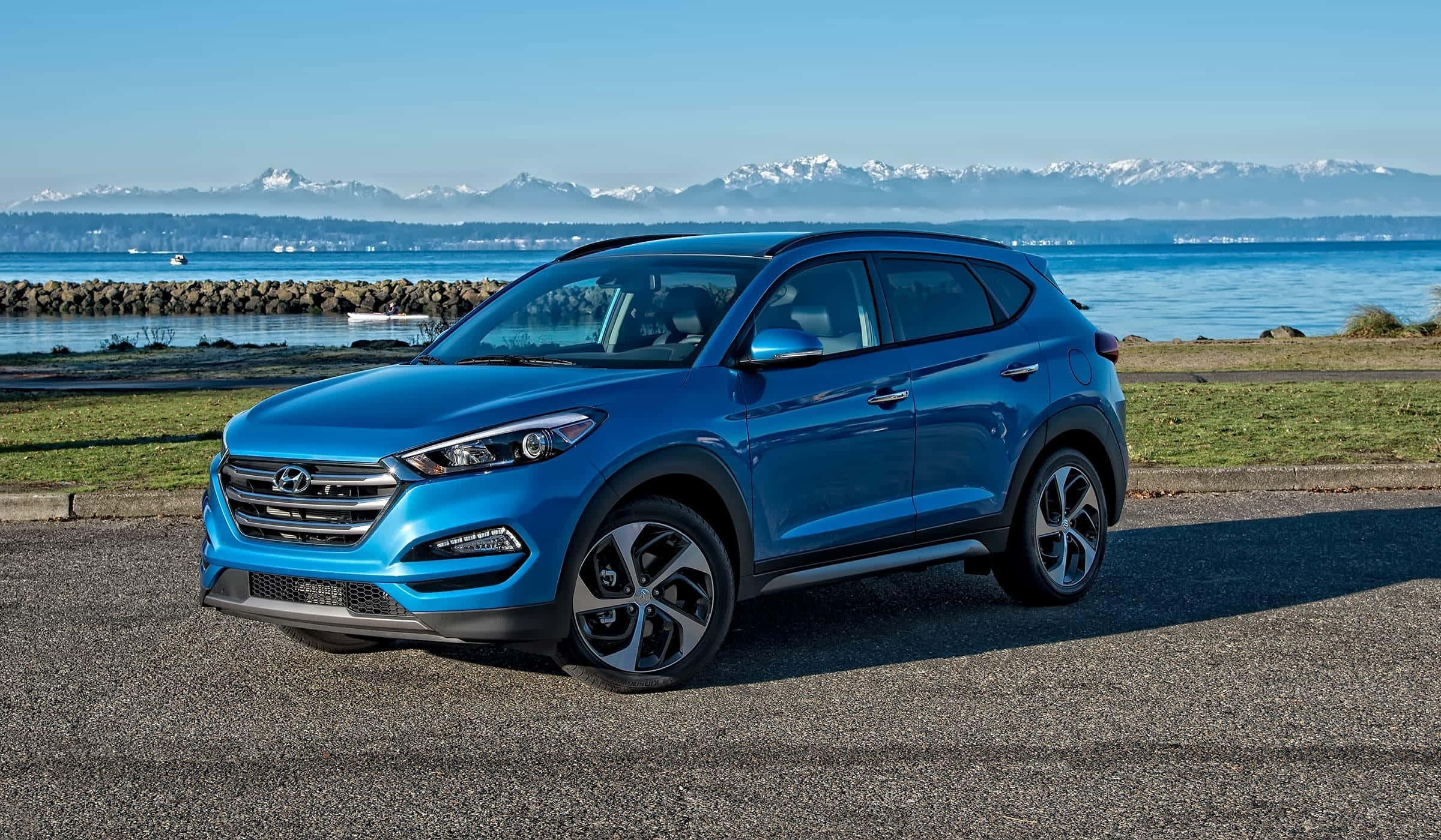 Hyundai Tucson 桌布 壁紙 — 一輛鮮藍色的現代Tucson SUV停在碎石地上，寧靜的海濱和遠處白雪覆蓋的山脈在清澈的藍天下。