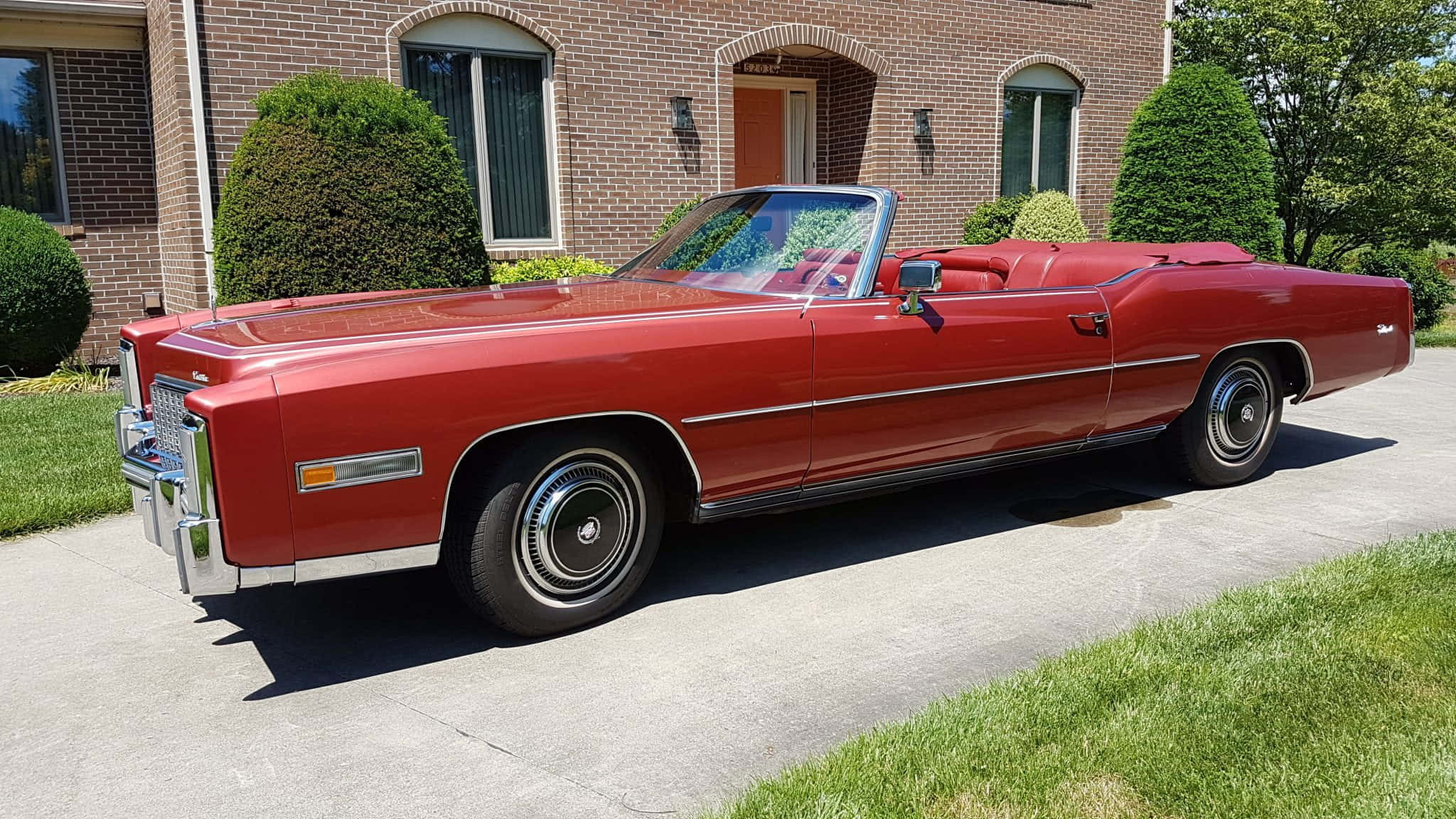 Cadillac Eldorado 桌布 壁紙 — 一輛光亮的紅色經典敞篷車停在磚房前的車道上，磚房周圍有修剪整齊的灌木叢。