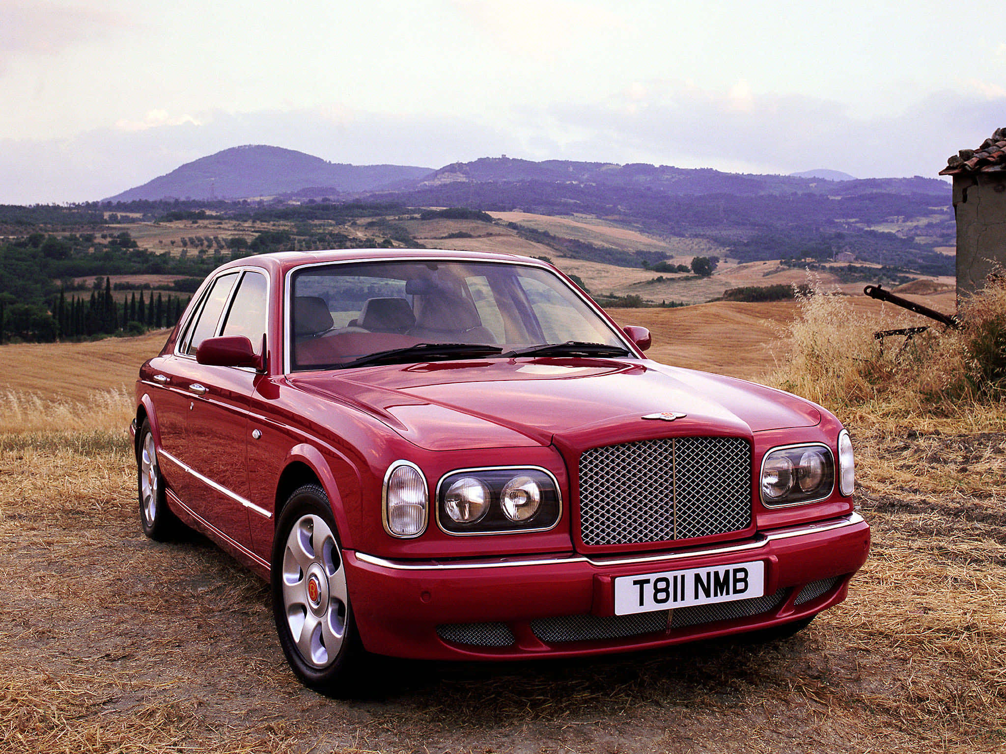Bentley Arnage 桌布 壁紙 — 一輛光亮的紅色賓利轎車停在乾草地上，背景有起伏的山丘和一座石建築，天空多雲。
