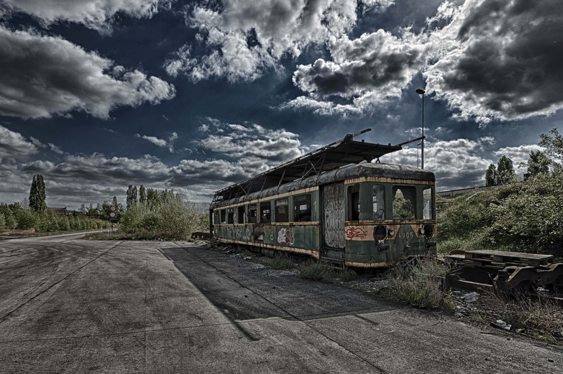  壁紙 — 一輛生鏽的廢棄火車車廂擱置在裂開的瀝青路面上，背景是戲劇性的暴風雨天空，周圍有野生植物攀爬。