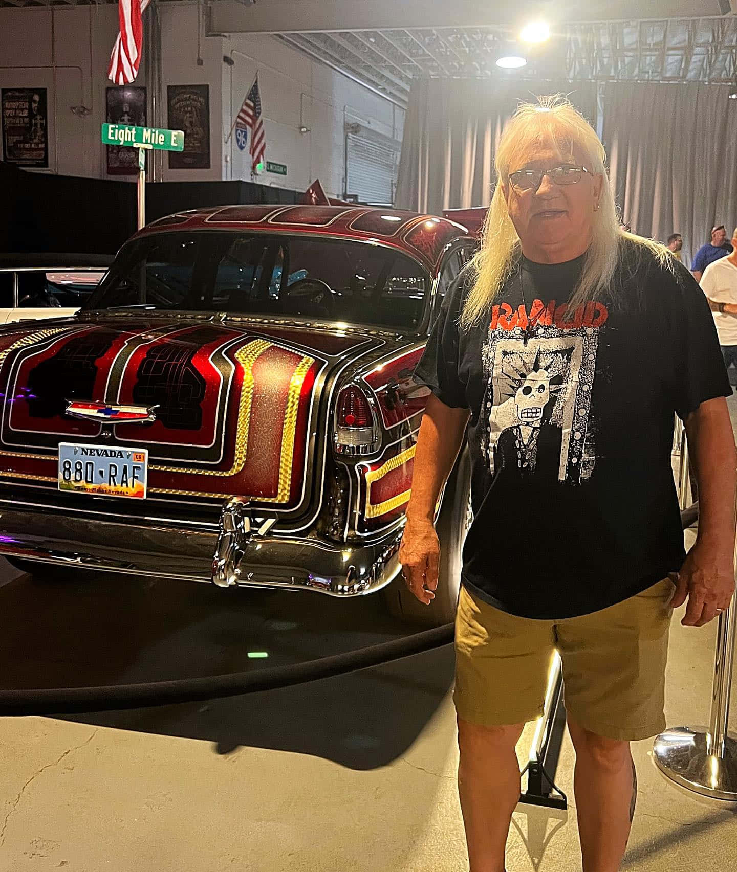  壁紙 — 一名長金髮男子站在明亮的燈光下，於室內車展中，旁邊是一輛訂製的紅金色改裝車。