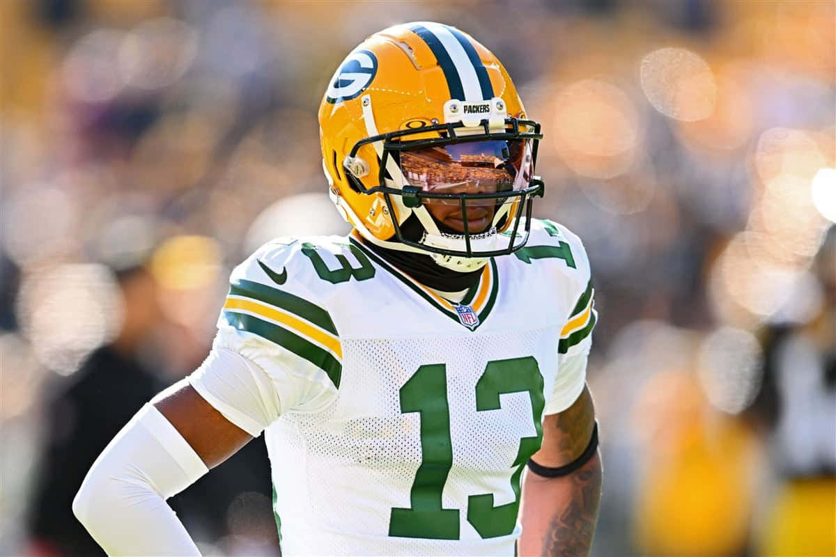  壁紙 — 一名身穿白色和綠色球衣、號碼13的綠灣包裝工美式足球員在明亮的日間比賽中專注地站在球場上。