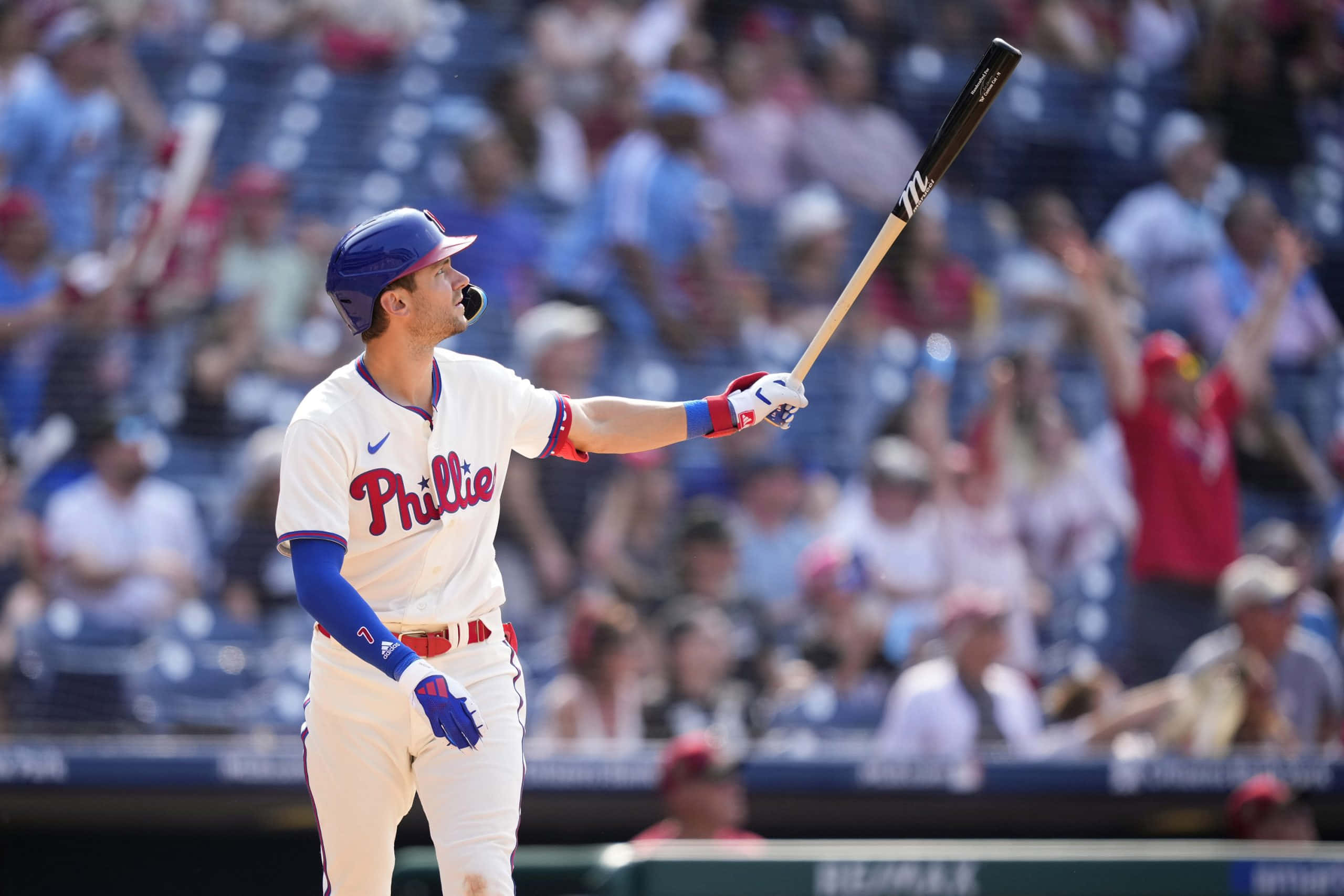 Trea Turner Phillies 桌布 壁紙 — 一名身穿白色球衣的費城費城人棒球選手，在陽光明媚的球場上，高舉球棒向歡呼的球迷致意。