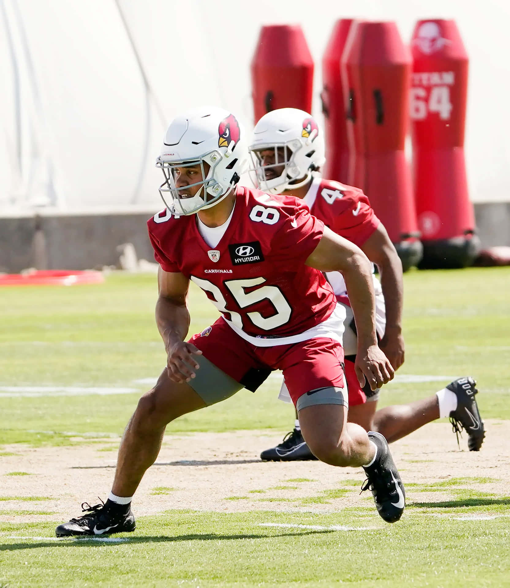  壁紙 — 一名身穿紅色 Cardinals 裝備的美式足球員在陽光明媚的場地上練習衝刺，背景有隊友和紅色的擒抱練習假人。