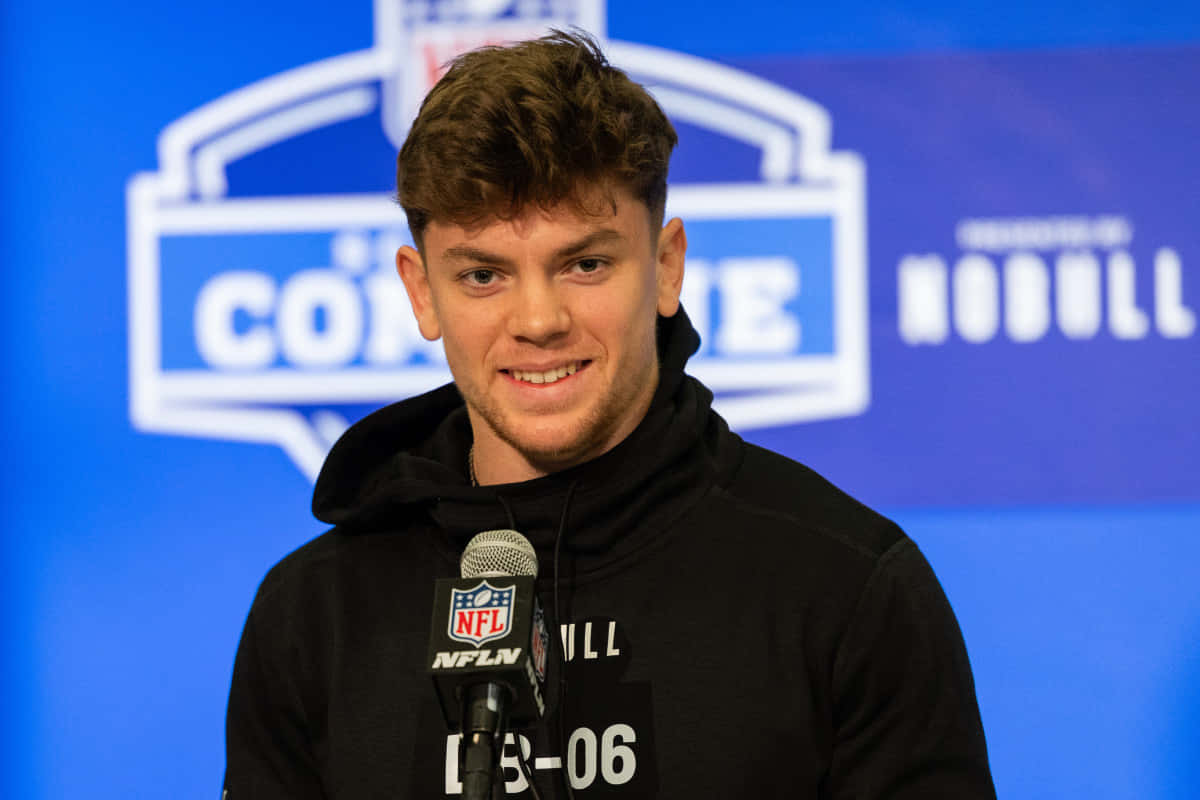  壁紙 — 一名穿著黑色連帽衫的年輕男子在NFL聯合測試記者會上，對著藍色品牌背景的麥克風微笑。