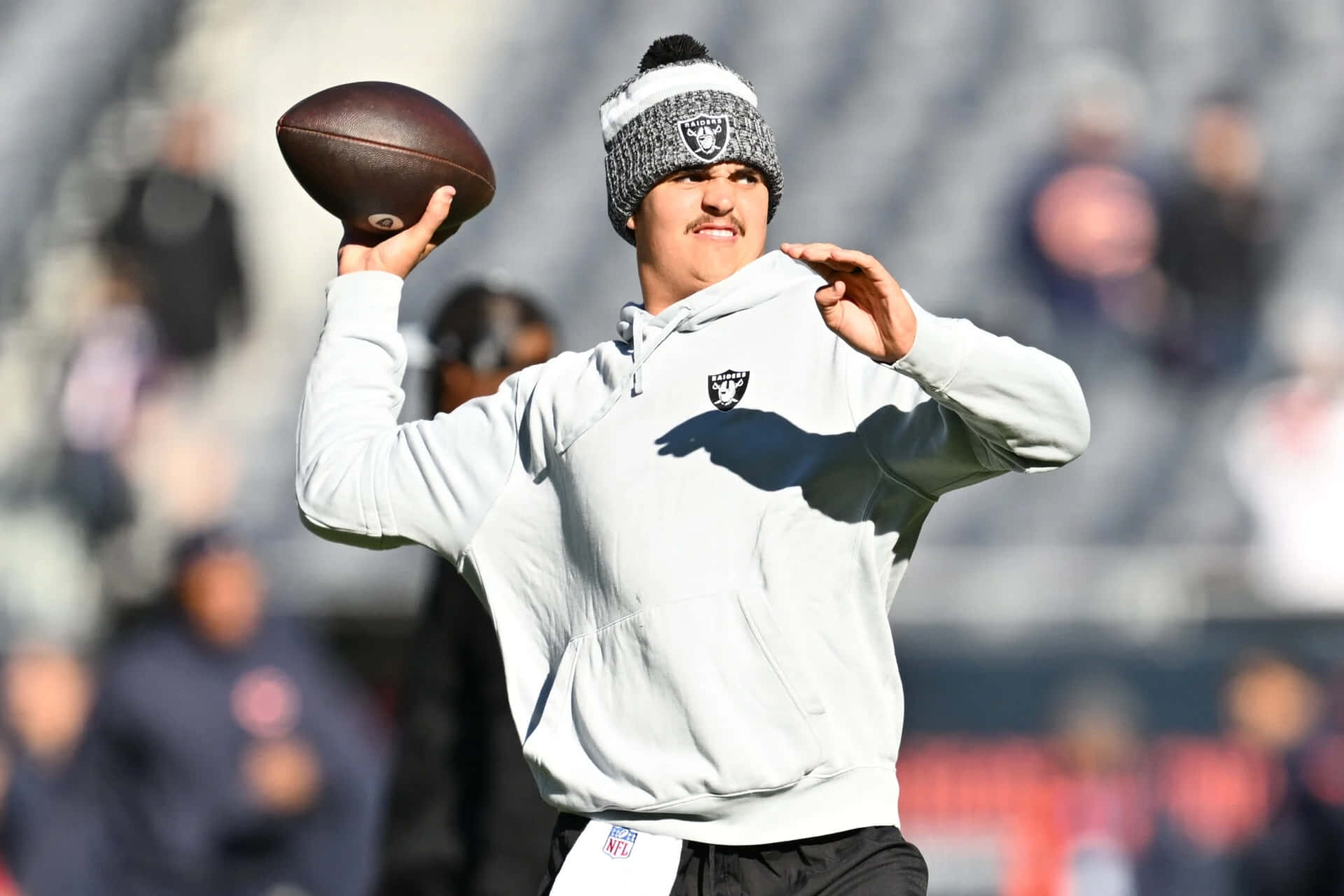  壁紙 — 一名穿著Raiders連帽衫和無邊帽的美式足球員在陽光明媚的日子裡在體育場準備投球。