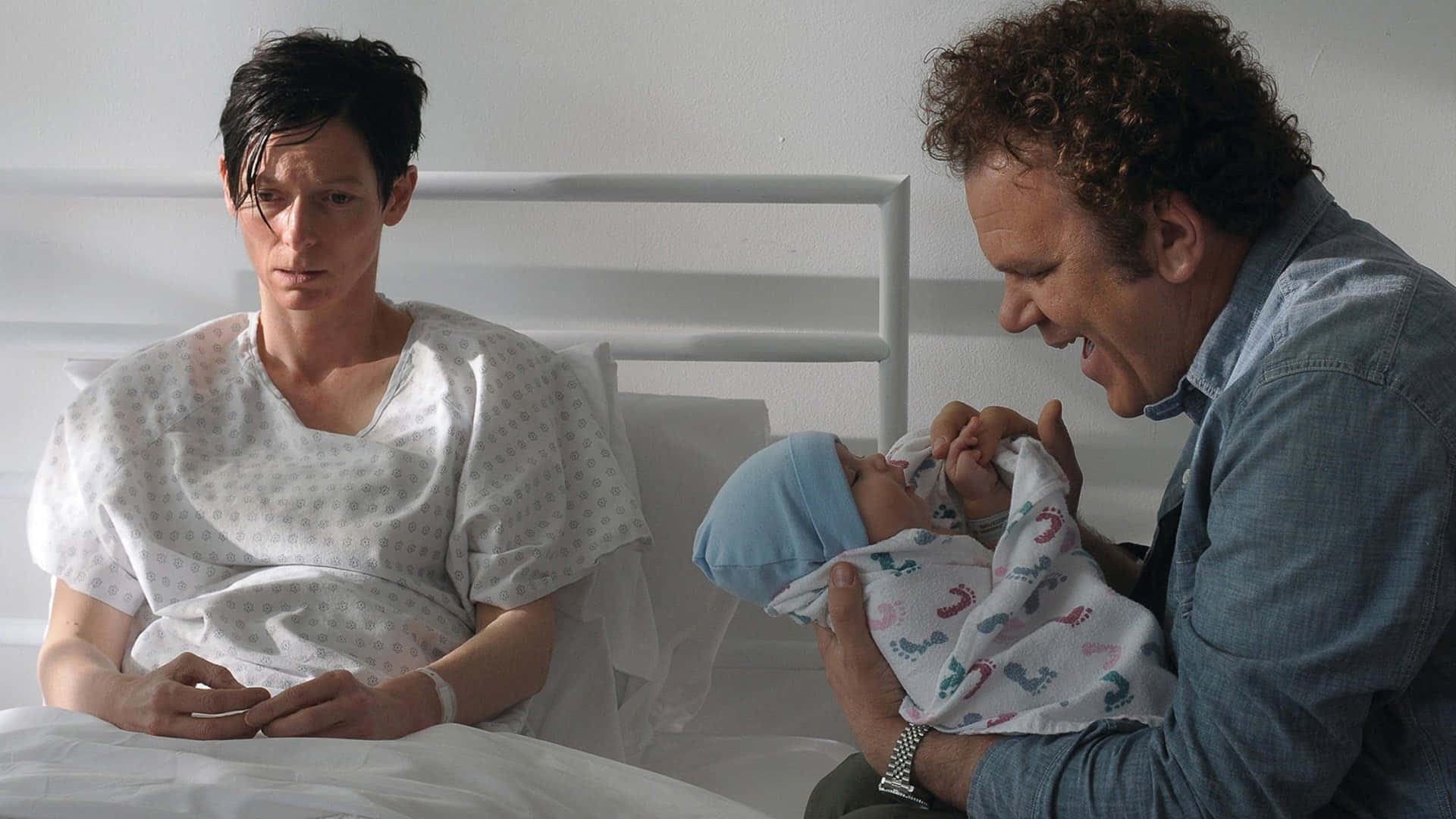 John C. Reilly 桌布 壁紙 — 一名穿著醫院袍的男人坐在床邊，另一名男人抱著一個用圖案毛毯包裹的新生嬰兒。